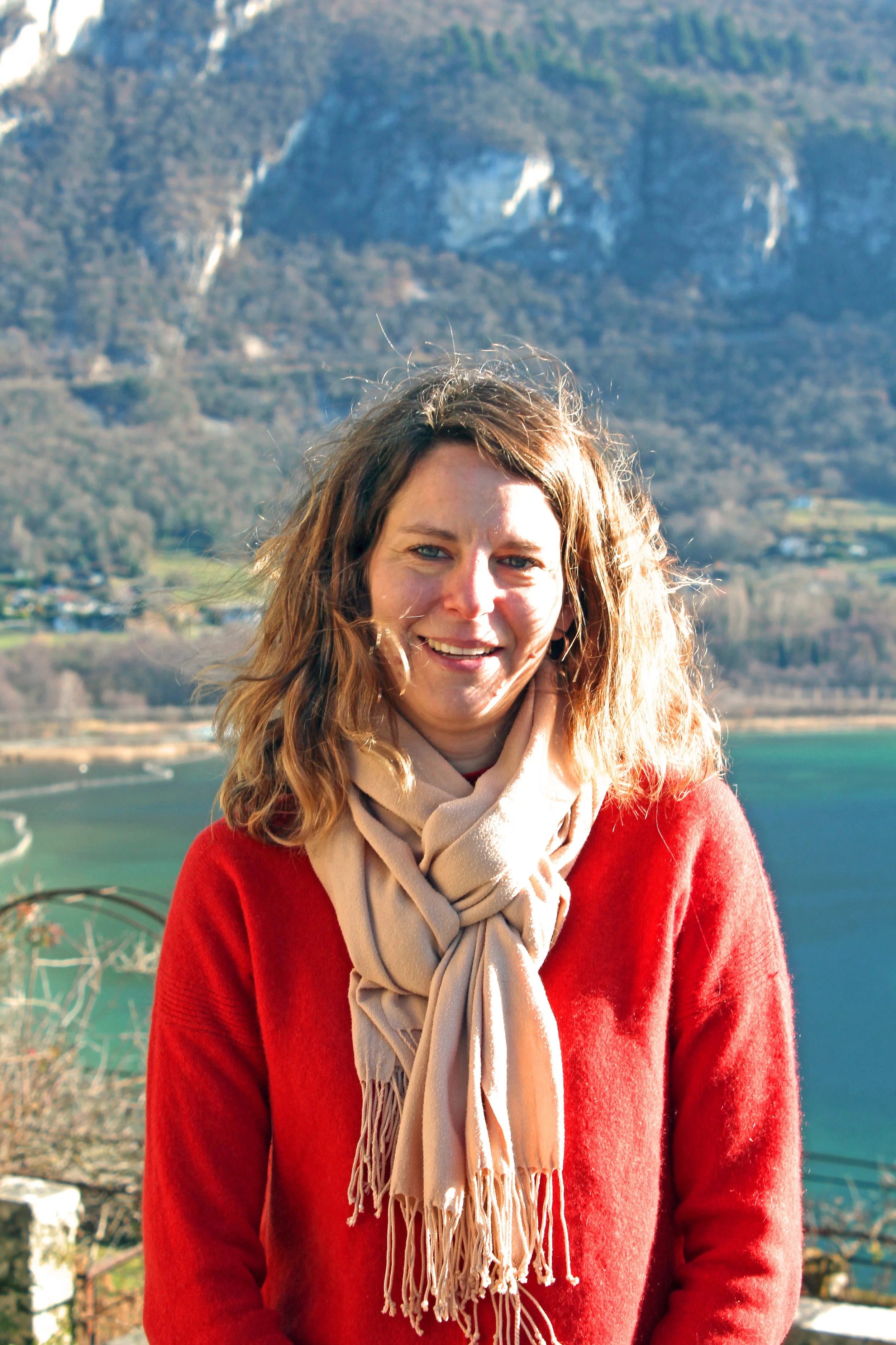 Une femme de Chindrieux souriante avec des cheveux ondulés bruns, portant un pull rouge et une écharpe beige, devant un paysage de montagnes et un lac.