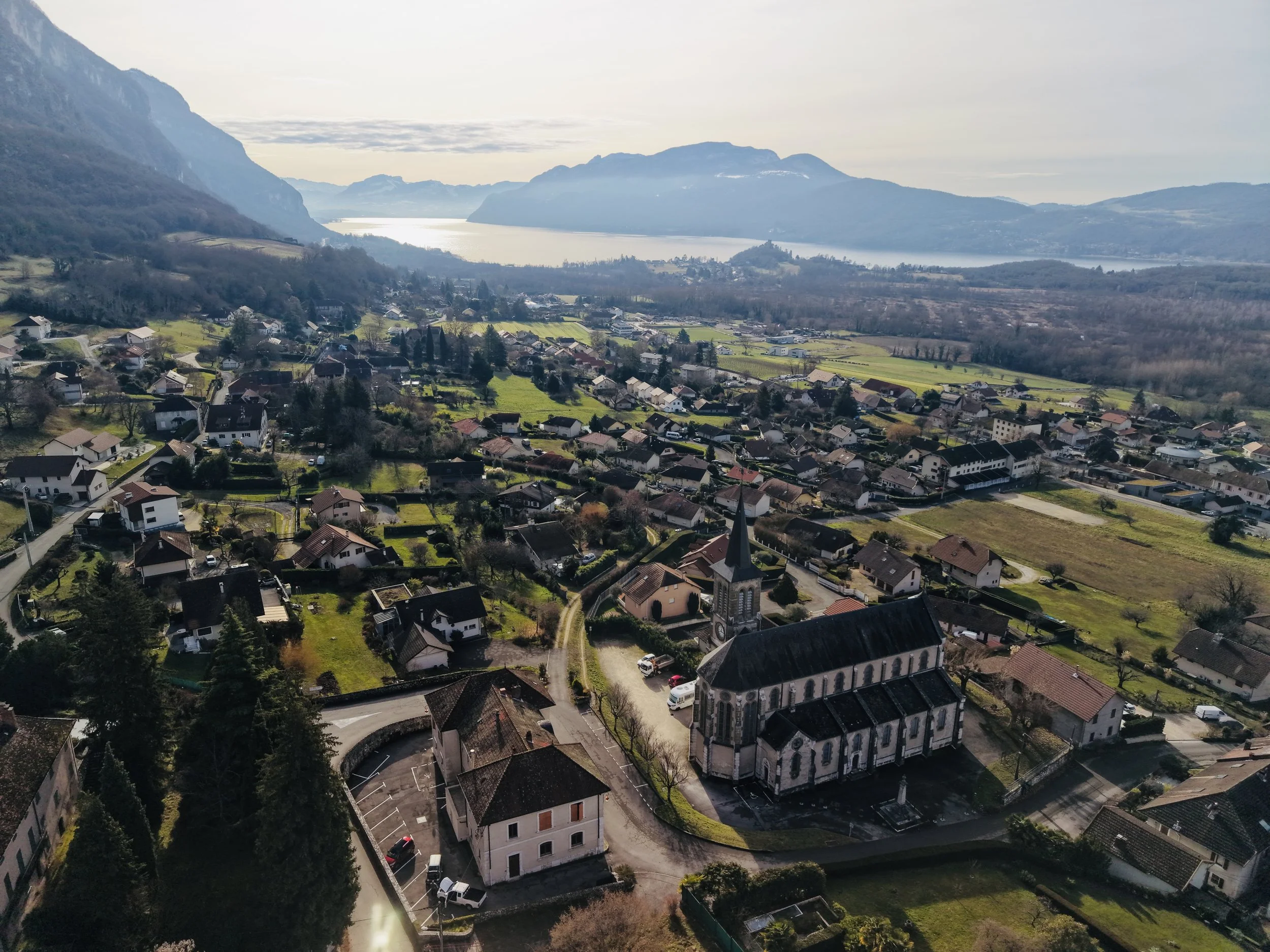 Vue aérienne d'un petit village Chindrieux avec une grande église au centre, entouré de maisons, de champs verts, avec un lac et des montagnes en arrière-plan.