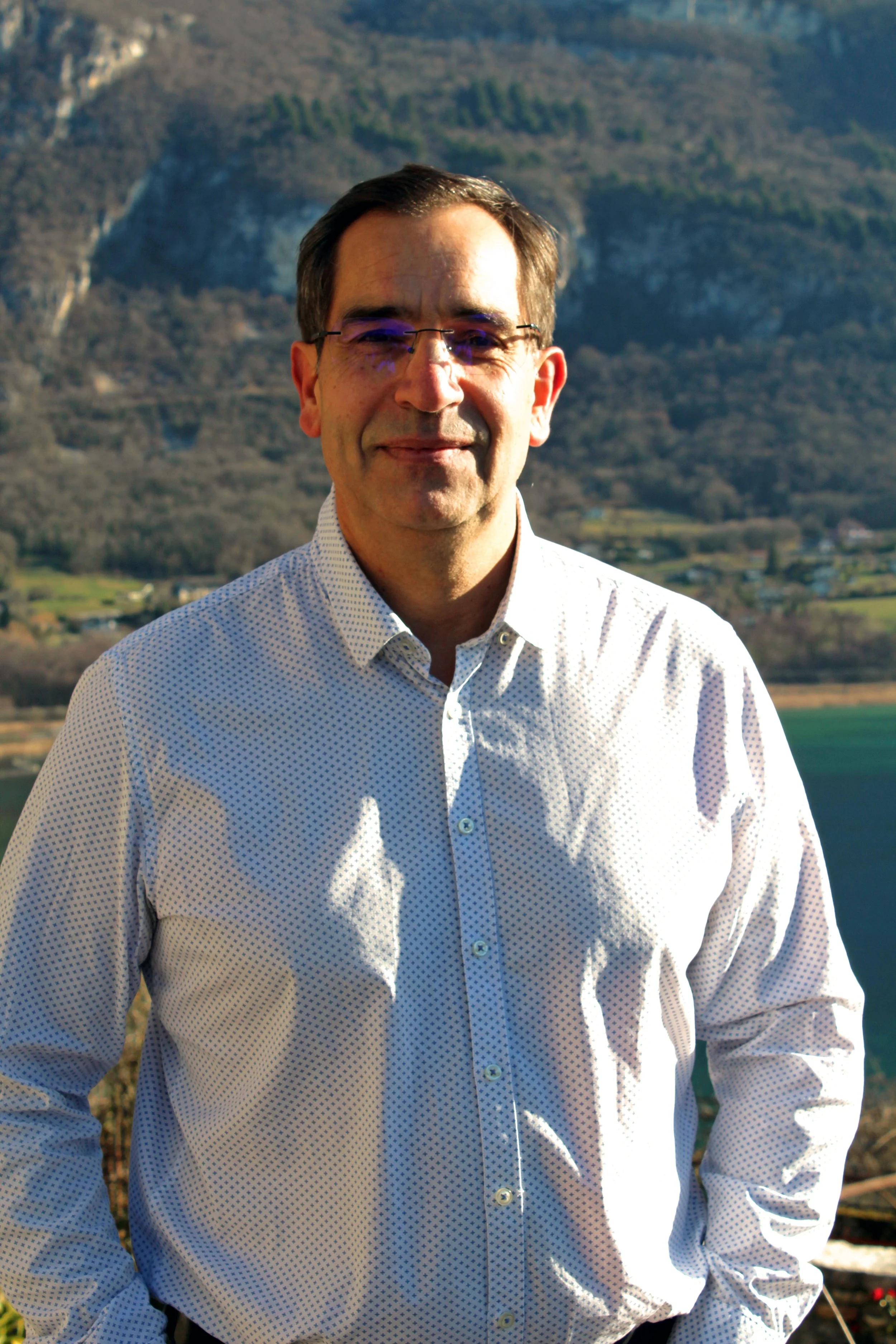 Un homme Chindrieux souriant porte une chemise à petits motifs blancs et bleus, debout devant un paysage de montagnes et de lac, en plein jour.