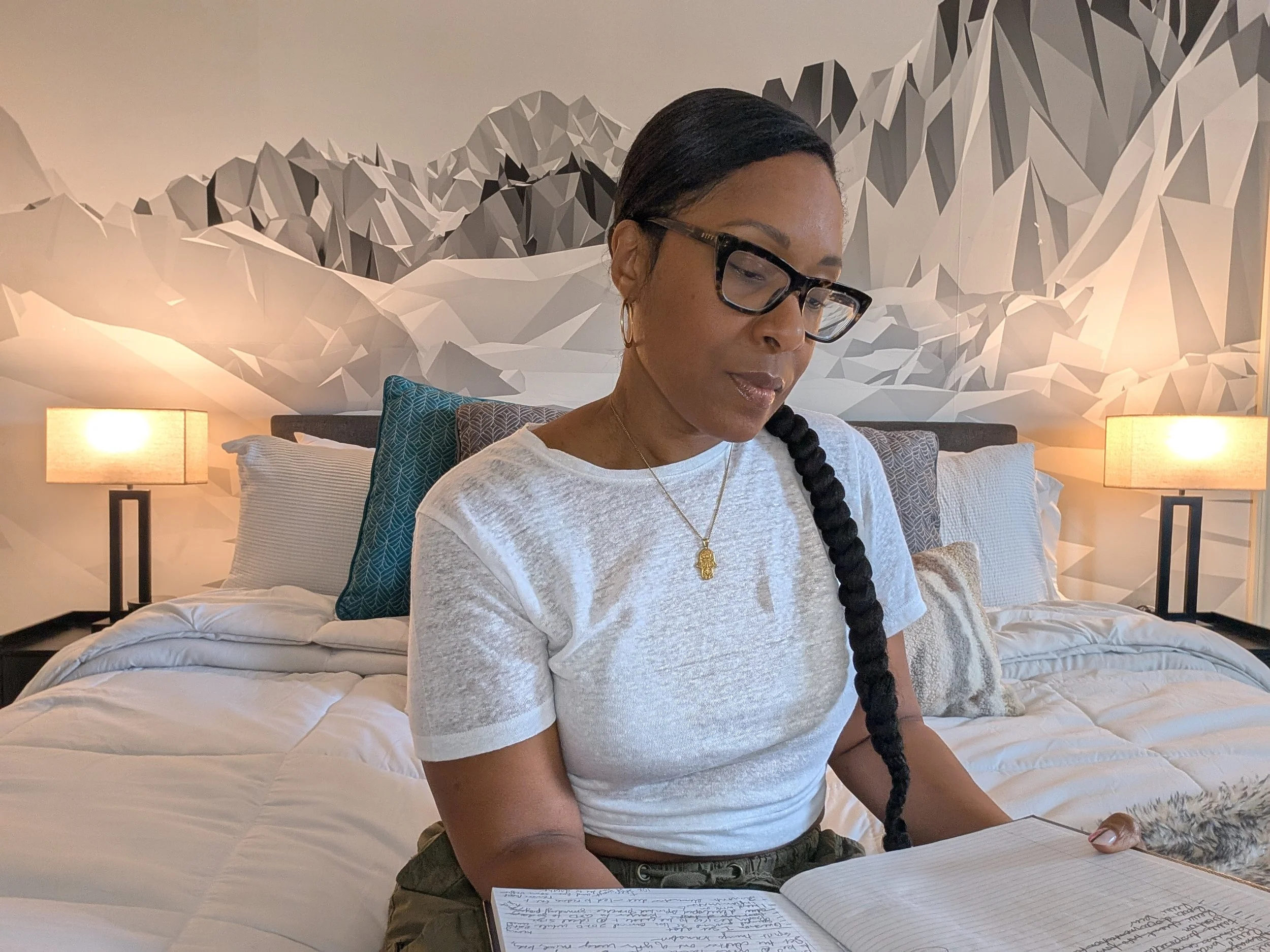 A woman with glasses and a long braid is sitting on a bed reading a notebook in a bedroom with modern mountain-themed wall art and two bedside lamps.
