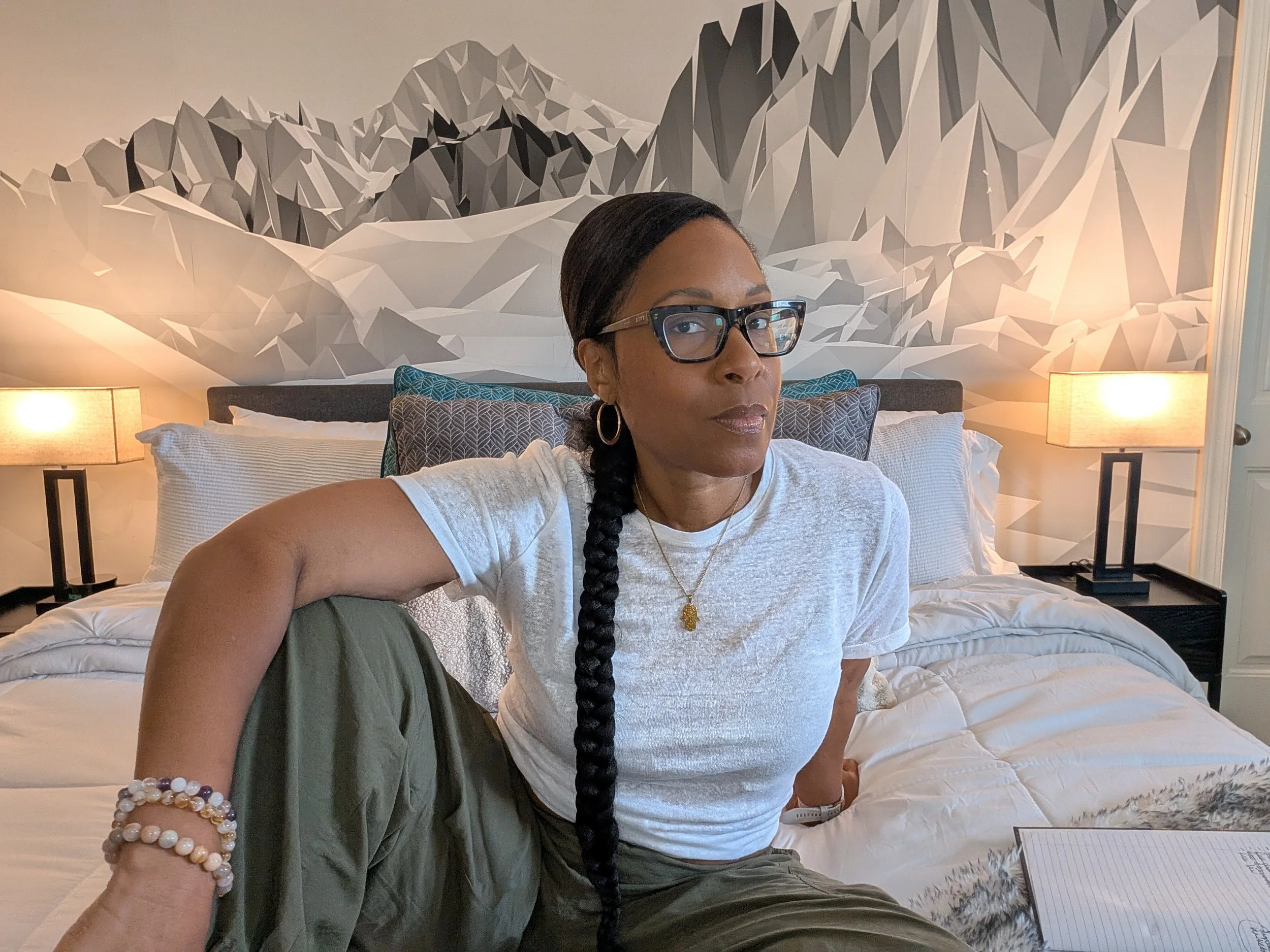 A woman with glasses and long black braid sitting on a bed with a mountain mural behind her, flanked by two lamps.