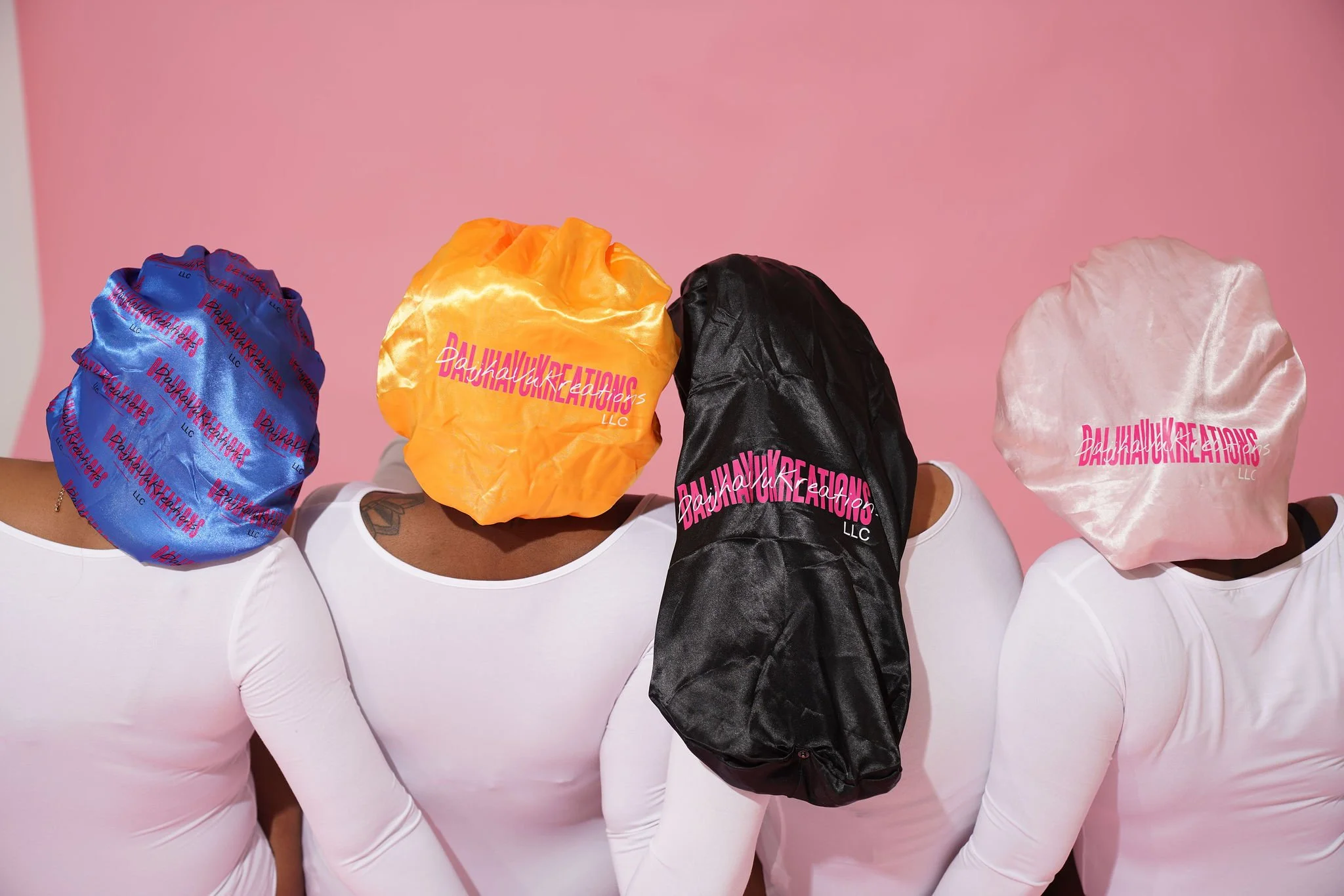 Four individuals sitting with their backs to the camera, each with a colorful satin bonnet on their head. The bonnets are blue, yellow, black, and pink, each branded with 'Baby Hawk Creations LLC' in pink text. The background is pink and they are all wearing white long-sleeve tops.