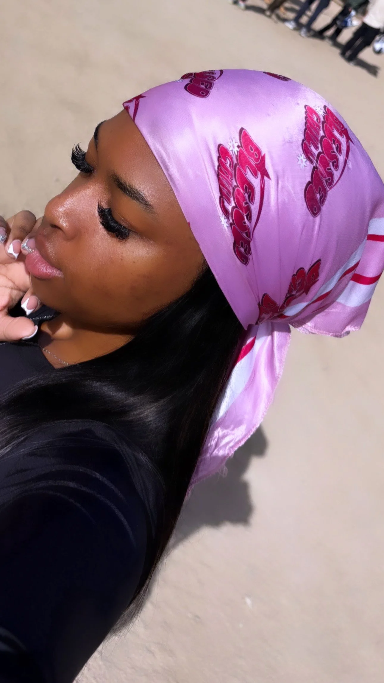 A young woman with long black hair wearing a pink satin bandana with red and white lettering that reads 'Cherry B'. She has long eyelashes, glossy lips, and is wearing a black top, standing outdoors.