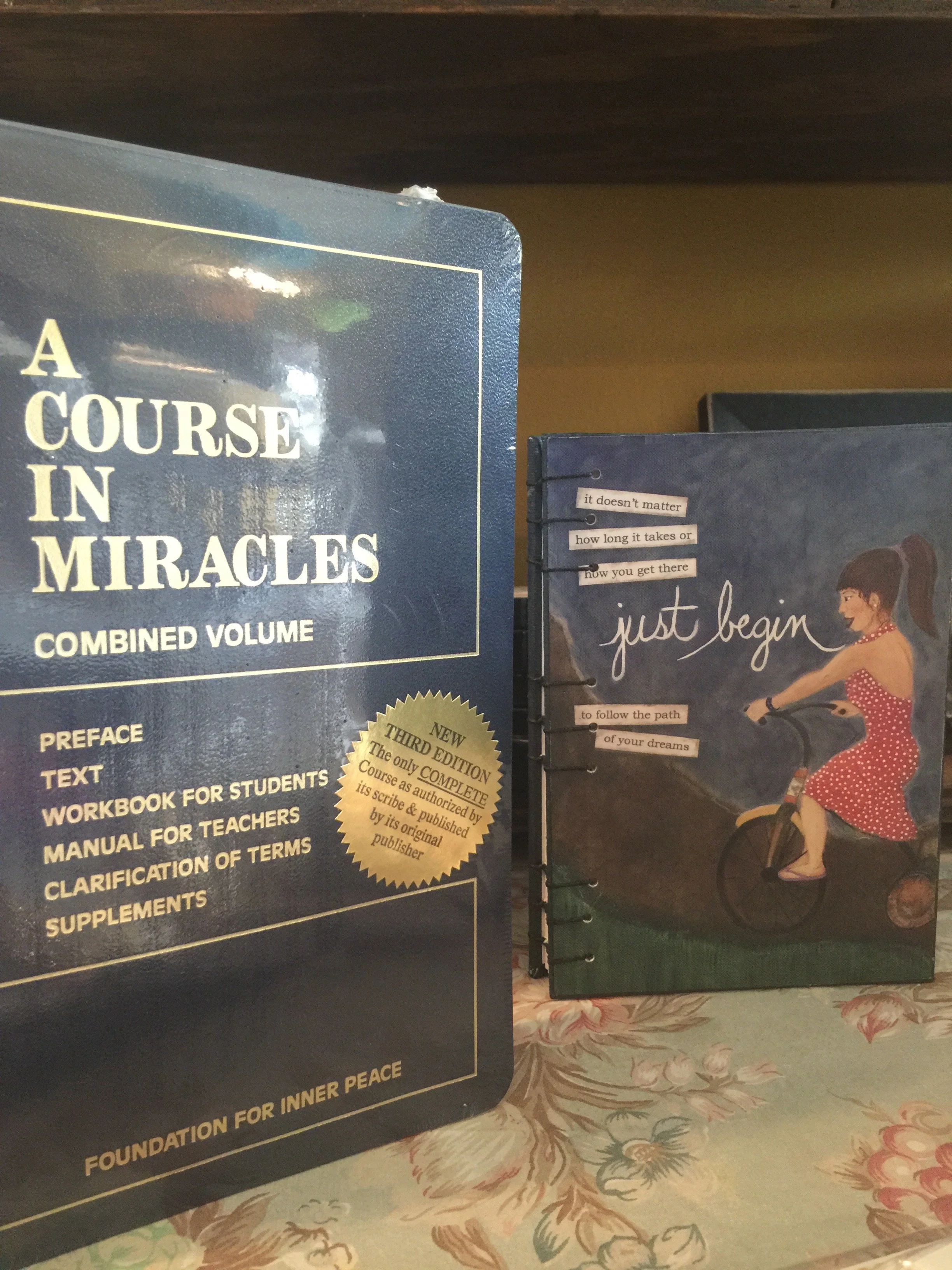 Close-up of two books on a table, one titled "A Course in Miracles" and the other with a cover illustration of a woman in a polka-dot dress riding a bicycle, with a background of a warm-colored wall and floral tablecloth.