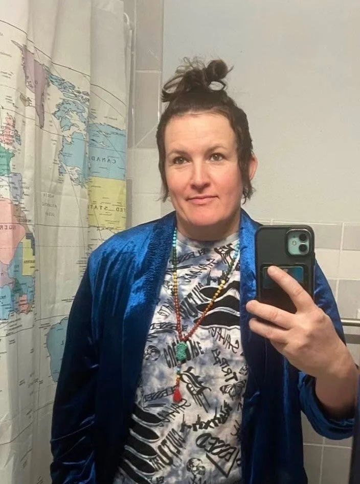 A woman taking a selfie in a bathroom mirror. She has short brown hair tied in a top knot, wearing a graphic T-shirt with black and white designs, layered necklaces, and a shiny blue jacket. The background features a bathroom shower curtain with a world map design.