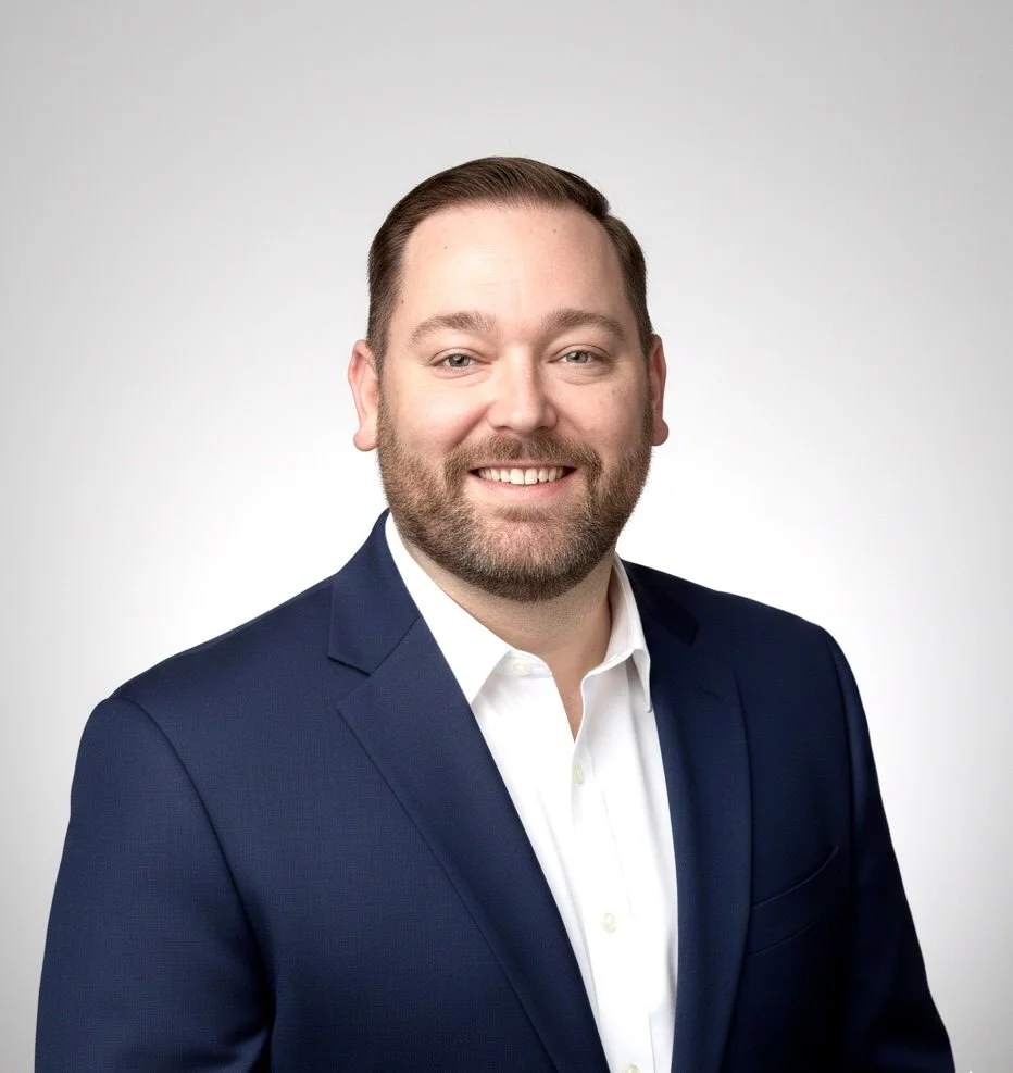 A professional headshot of a smiling man with short brown hair and a beard, wearing a navy blue suit and white dress shirt, against a plain white background.