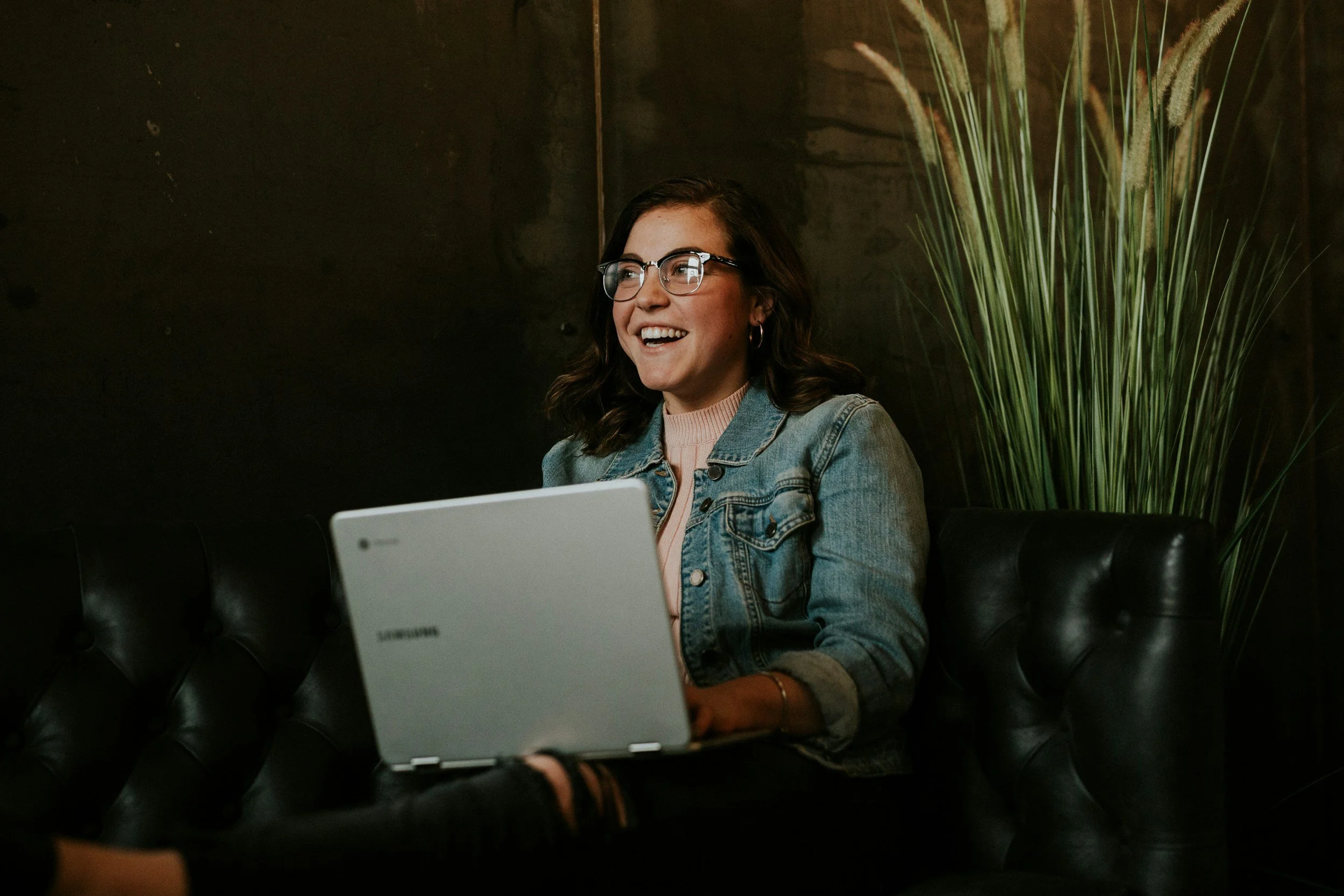 A young woman with glasses and earrings sitting on a black leather couch, smiling at a laptop, with teal walls and decorative tall green plants in the background.
