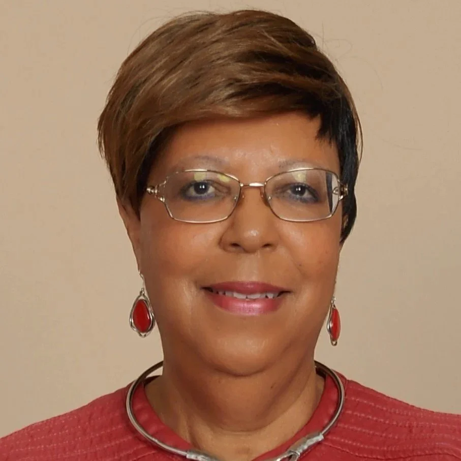 A woman with short brown hair, glasses, and earrings, smiling at the camera. She is wearing a red top with a matching necklace and earrings, with a neutral background.