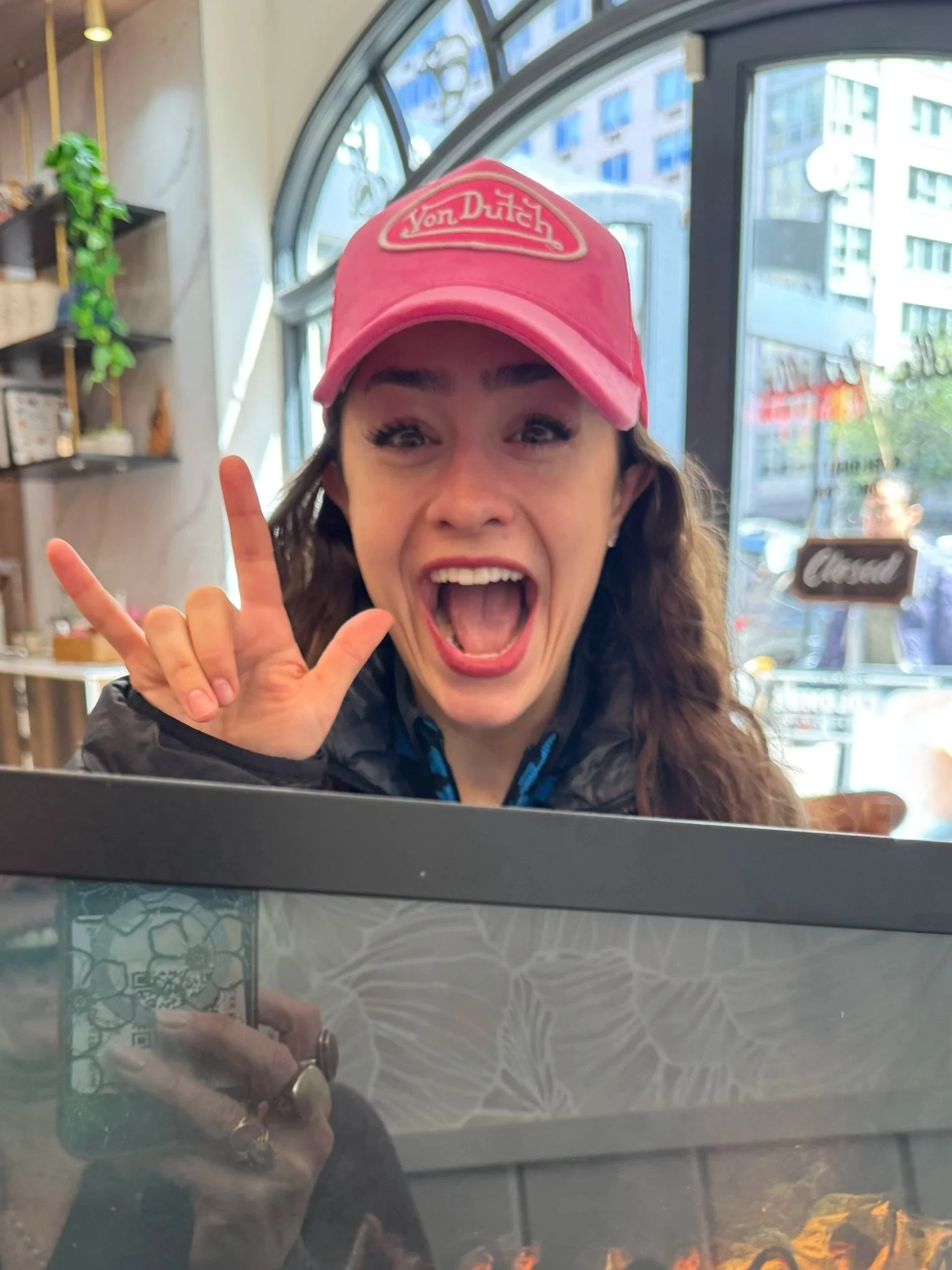 A woman wearing a pink Von Dutch cap, smiling widely and making a rock and roll hand gesture while inside a cafe or restaurant with large windows showing city buildings outside.