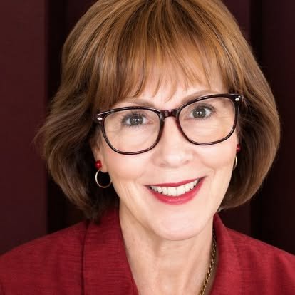 A woman with short brown hair, wearing glasses, red lipstick, and a red blazer, smiling at the camera.