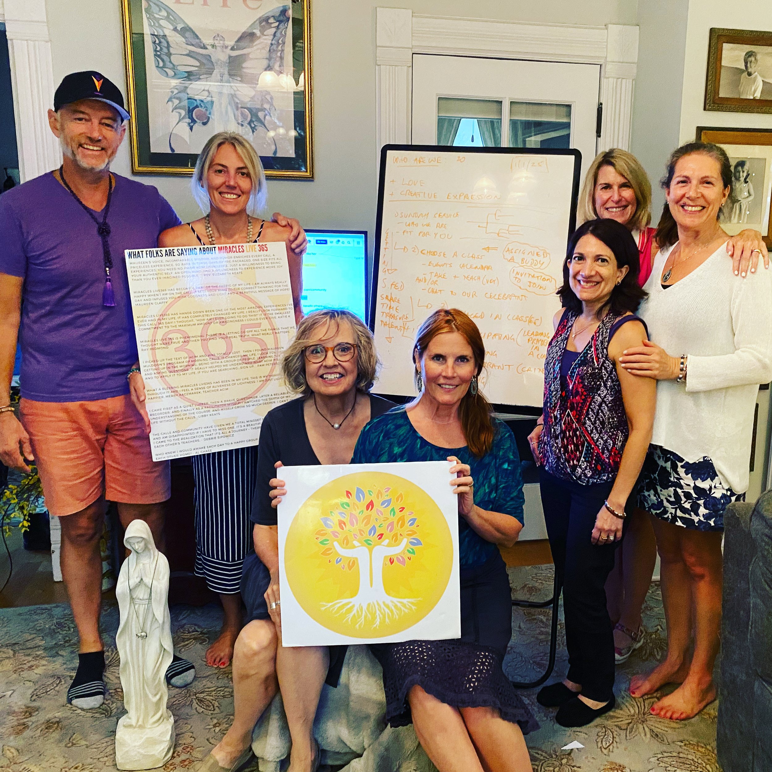 Group of six women and one man posing in front of a whiteboard with orange handwritten notes and a colorful tree graphic. Man on the left is wearing a purple T-shirt, orange shorts, black and white striped socks, and a black hat. The women are dressed in various colorful outfits, some holding signs or posters, and smiling at the camera. The setting appears to be a conference or workshop room with framed artwork on the walls.