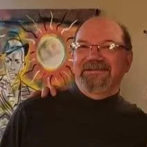 A man with glasses and a beard smiling in front of a colorful wall art featuring a face and a sunburst design.