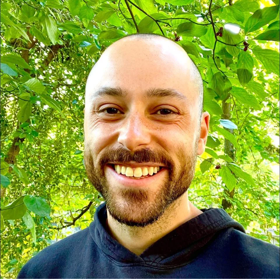 A smiling man with a beard and shaved head standing in front of green leaves in a natural outdoor setting.