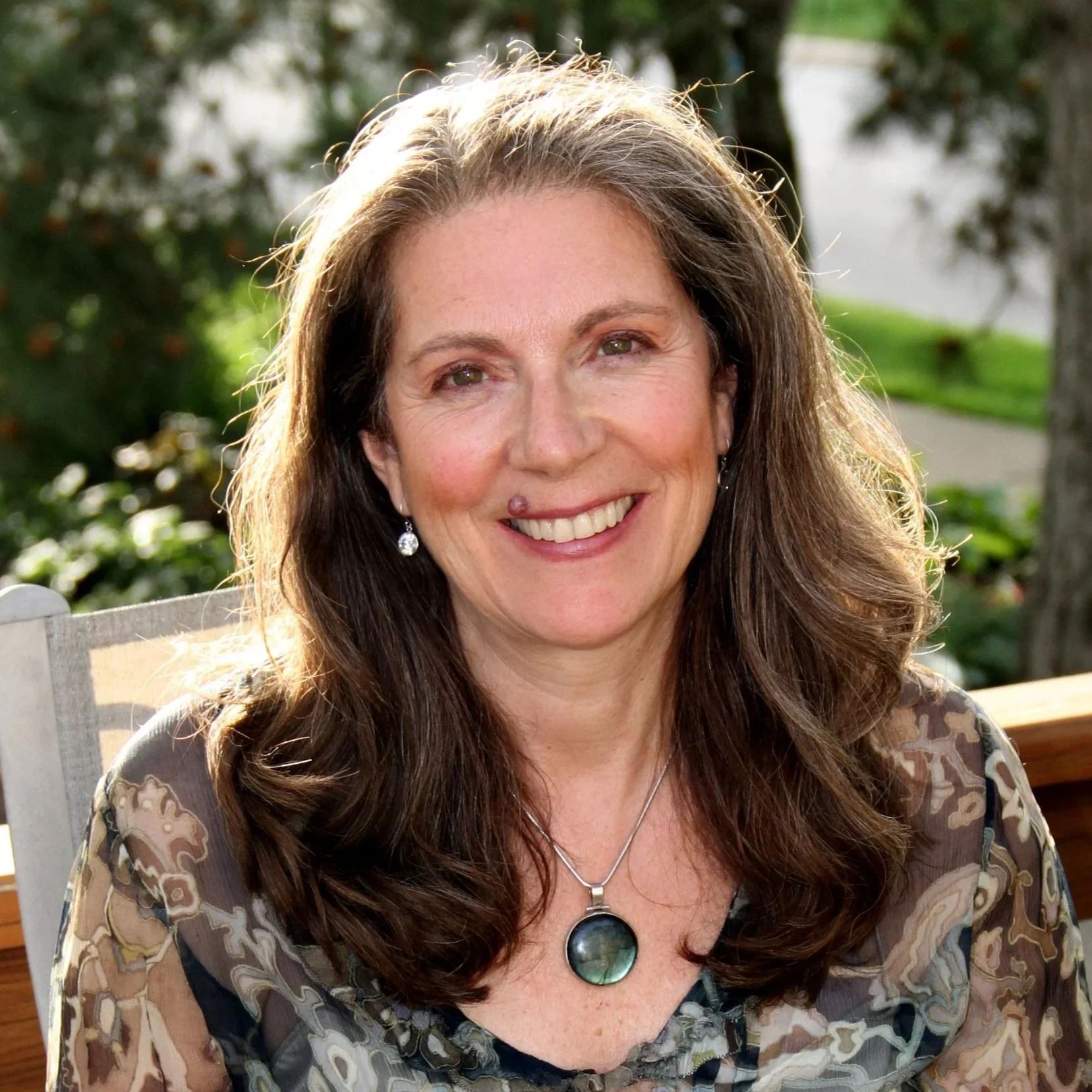 A woman with long brown curly hair smiling outdoors, wearing earrings and a pendant necklace, seated on a wooden bench with a blurred background of trees and greenery.