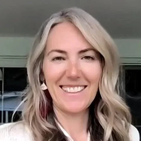 A woman with blonde, wavy hair smiling in a well-lit room.