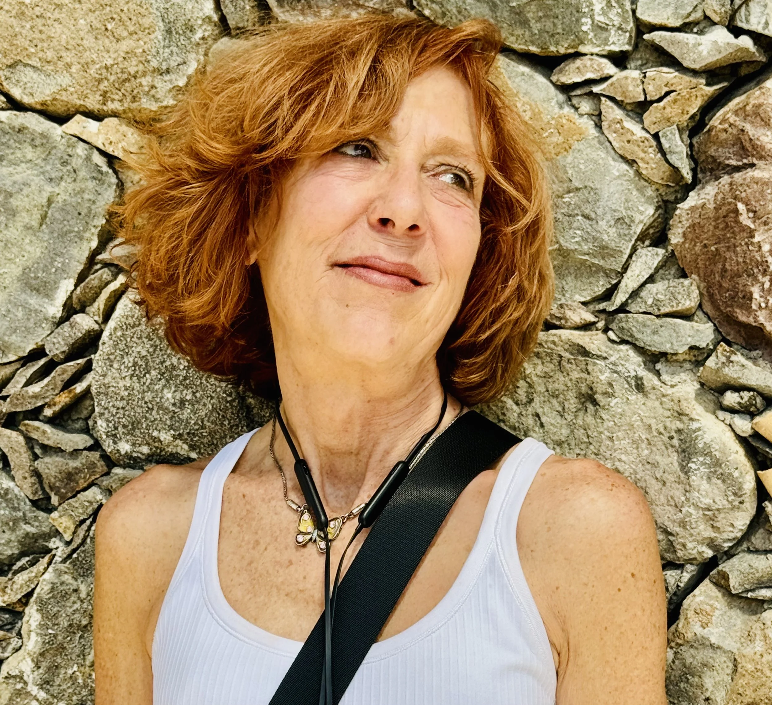 A woman with red hair lying against a stone wall, wearing a white tank top, with headphones around her neck, smiling and looking slightly to her left.