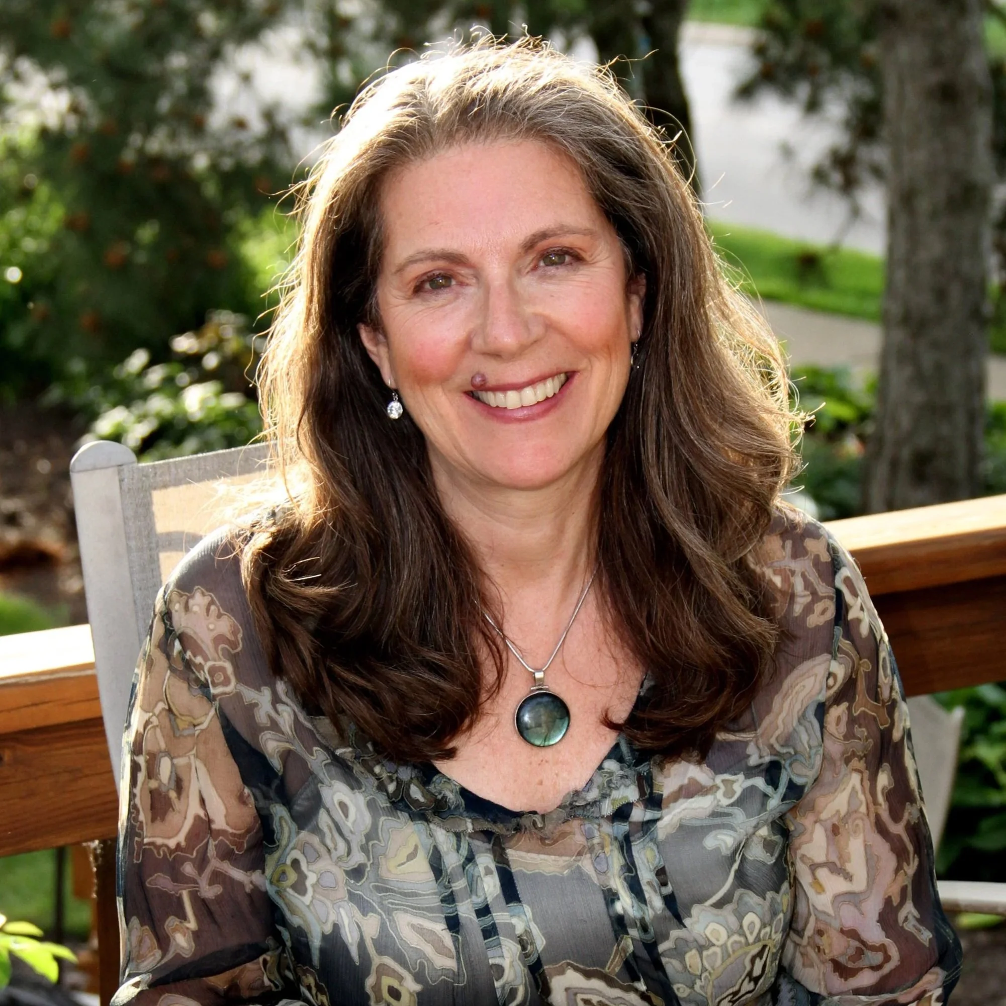 A middle-aged woman with long brown hair, wearing earrings and a pendant necklace, smiling outdoors with greenery in the background.