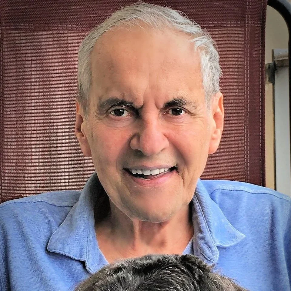 An older man with gray hair smiling, wearing a blue shirt, sitting in front of a red background, with part of an animal, possibly a dog, visible in the bottom of the image.