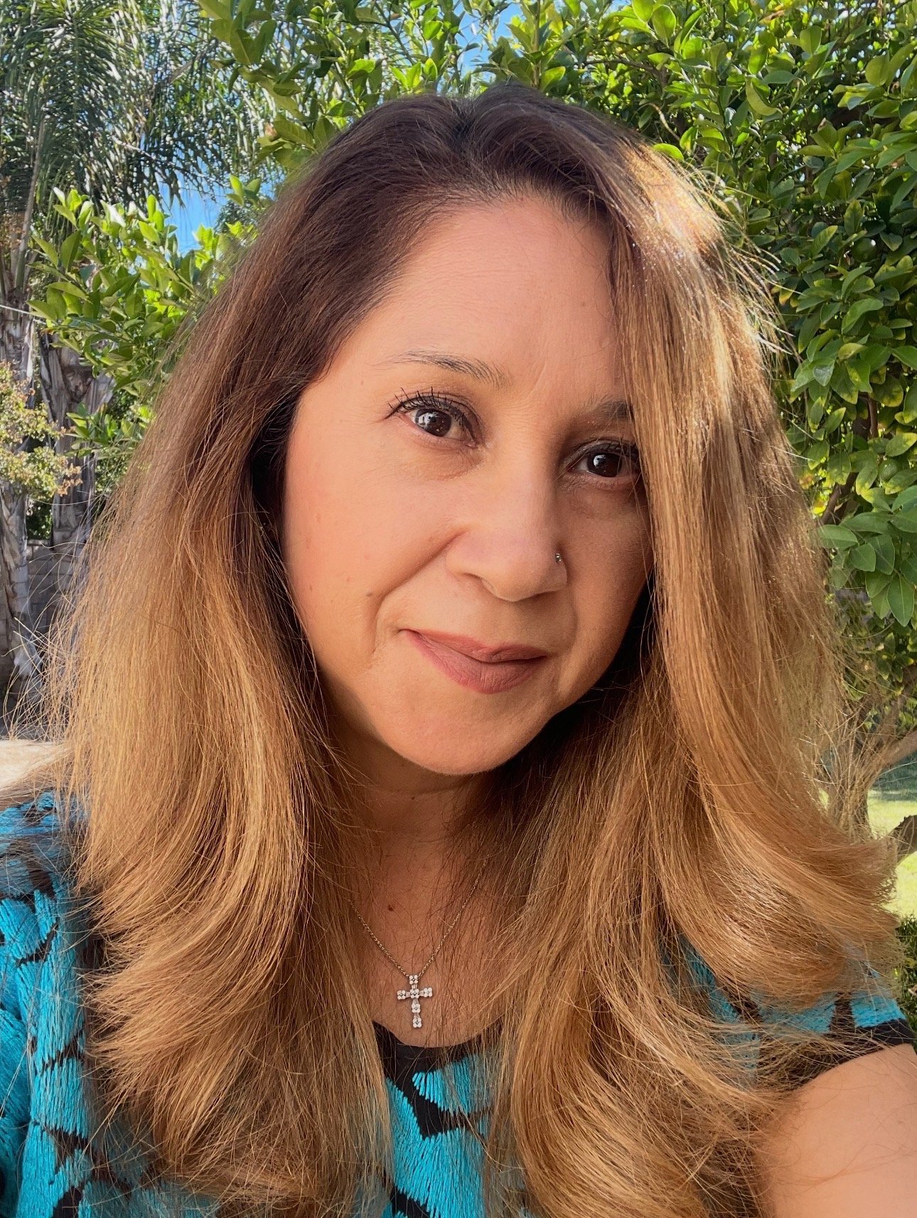 Close-up of a woman with long, wavy light brown hair, wearing a cross necklace and a turquoise and black patterned top, outdoors surrounded by green foliage.