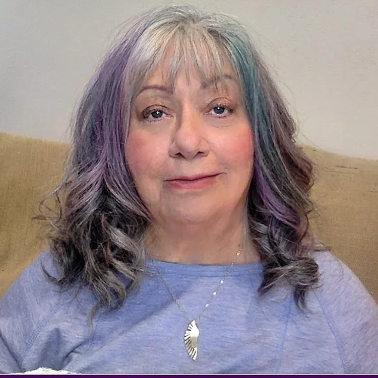 A middle-aged woman with gray and purple hair sitting on a beige couch, wearing a lavender shirt and a silver necklace with a feather pendant.