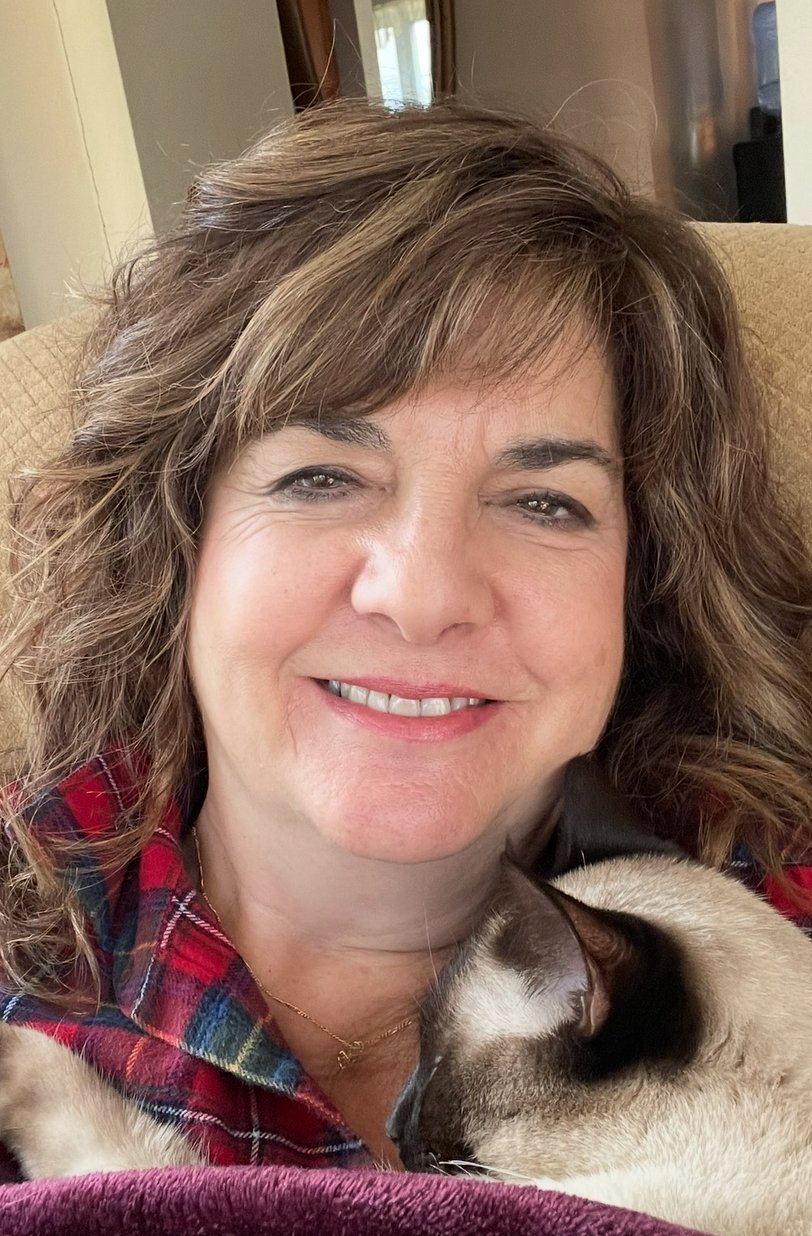 A woman smiling while sitting on a beige armchair, with a Siamese cat like breed resting on her chest.