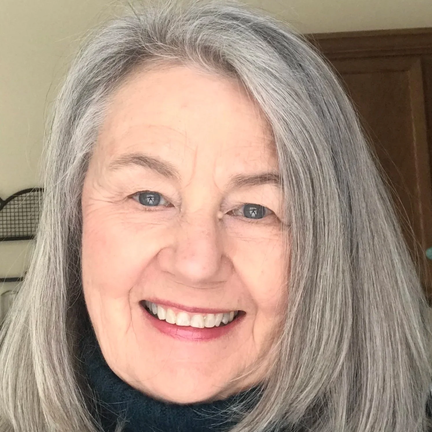 Close-up of a smiling woman with long gray hair and bright blue eyes in an indoor setting.