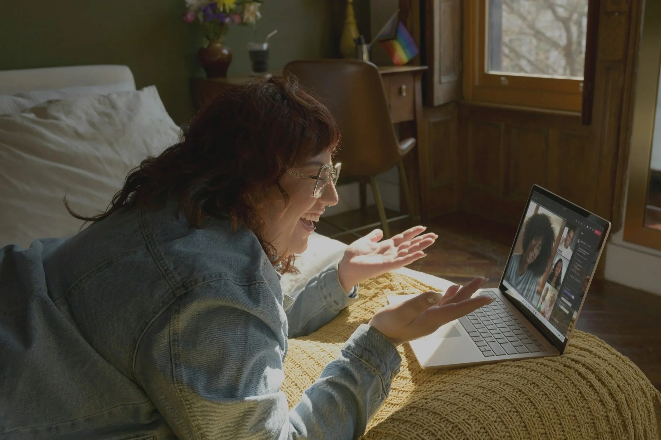 A woman with curly hair and glasses smiling and engaging via video call on a laptop, lying on a bed with a yellow blanket in a cozy room with a window and wooden furniture.