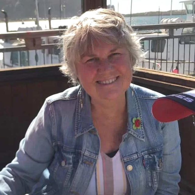 A smiling woman with short, wavy gray hair wearing a denim jacket with a flower embroidery on the collar, sitting outdoors near water and boats.
