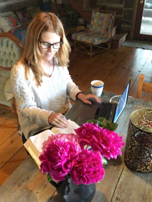A woman with glasses, wearing a sweater, is sitting at a wooden table with an open book, a laptop, a cup, and a vase of pink flowers in a cozy room.