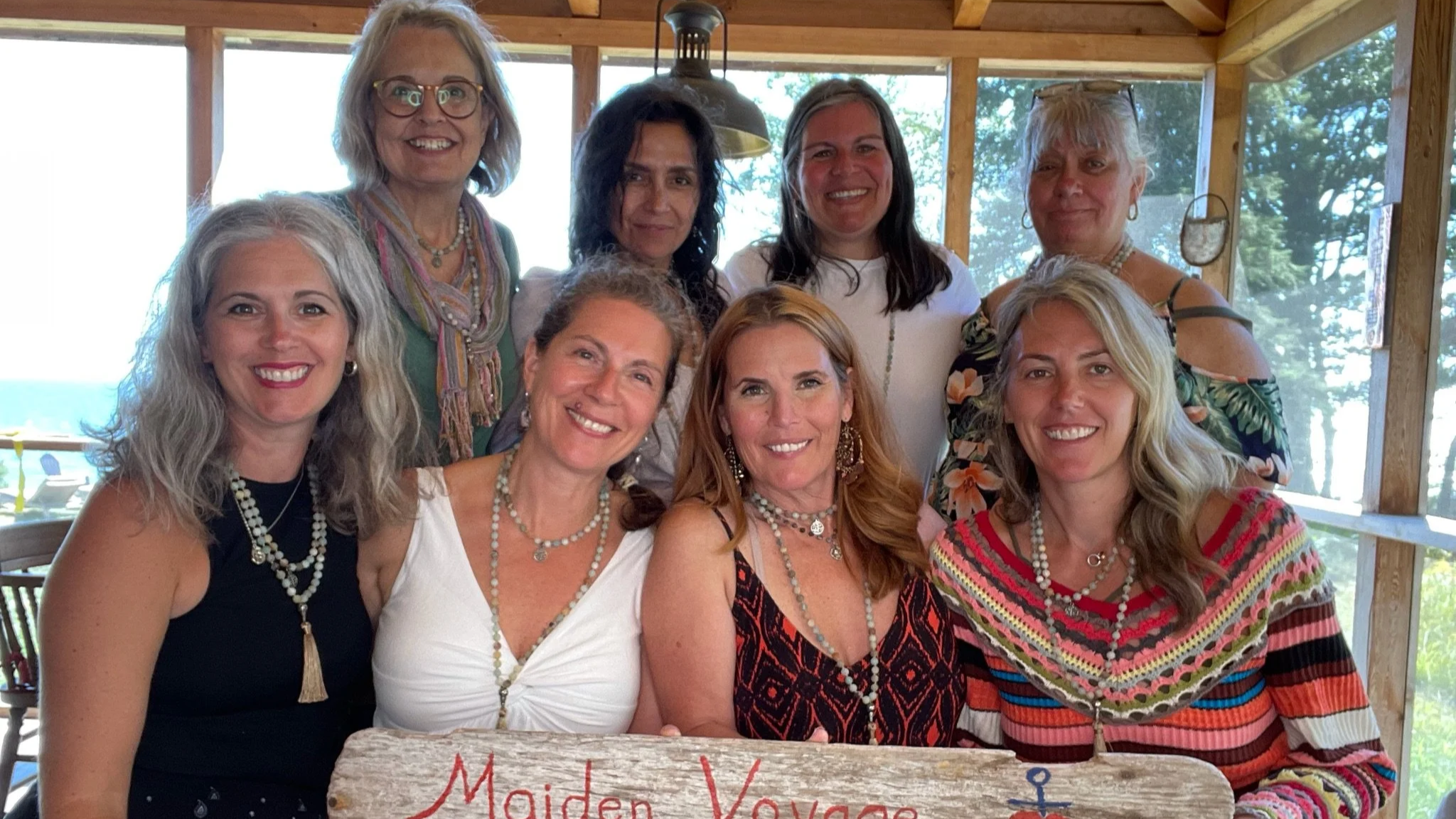 A group of ten women smiling and posing together indoors near large windows with trees visible outside. They are dressed in colorful and patterned summer clothing, and some are wearing jewelry.