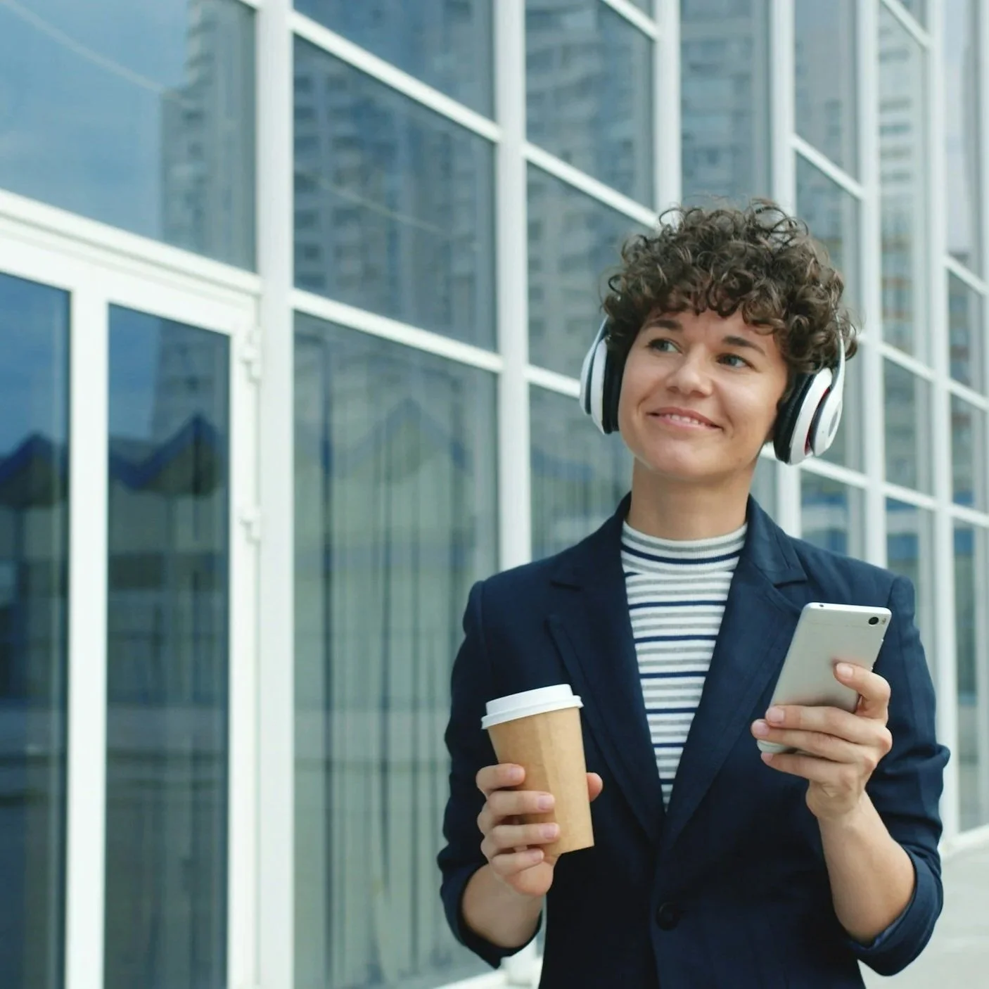 A woman with curly hair wearing headphones, a striped shirt, and a blazer, smiling while holding a smartphone in one hand and a coffee cup in the other, standing outside a modern building with glass windows.