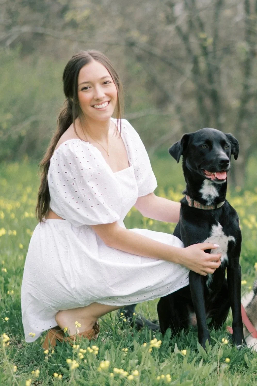 A woman in a white dress kneels on grass, smiling, with her hand on a black dog sitting next to her in a field with yellow flowers and trees in the background.