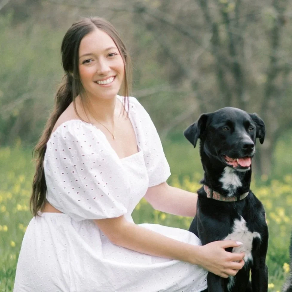 A young woman in a white dress sitting outdoors with a black dog.
