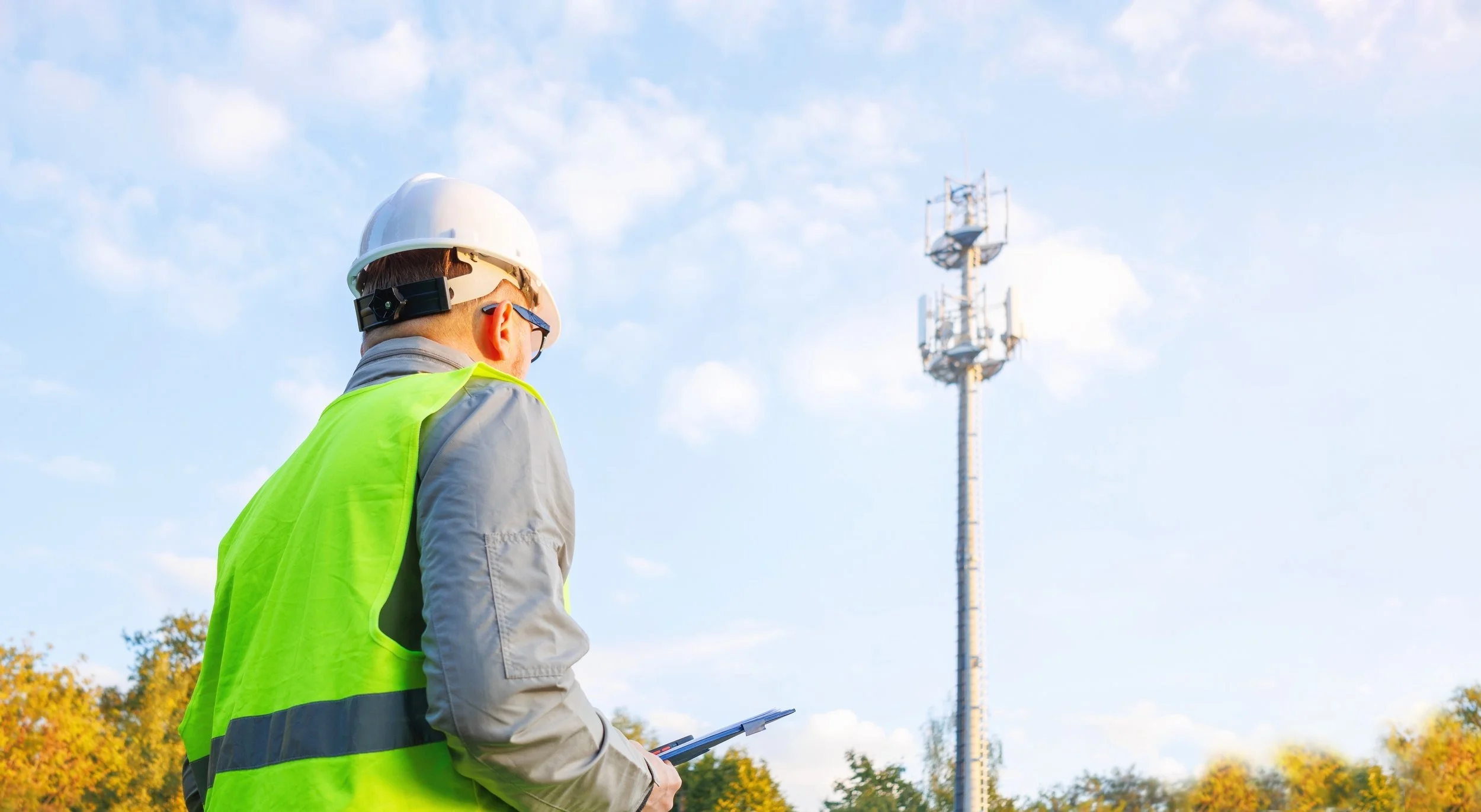 A man wearing a white safety helmet, sunglasses, and a reflective yellow vest, holding a clipboard, and looking at a tall communication tower against a blue sky with some clouds.