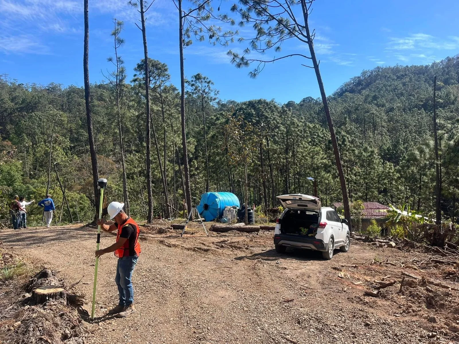 Construcción en un área forestal con varios técnicos trabajando, un coche con su maletero abierto, maquinaria y un tanque grande azul en un entorno de árboles y colinas.