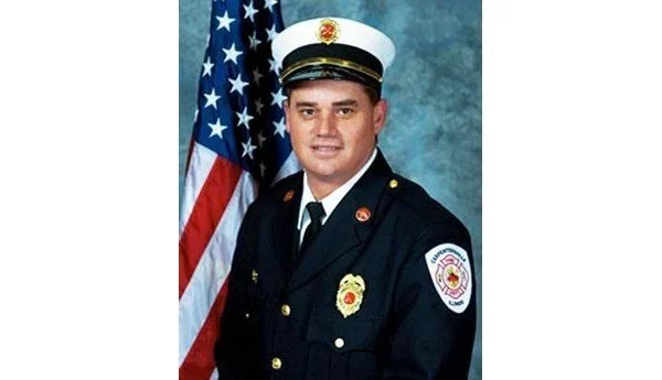 Formal portrait of Battalion Chief Todd Middendorf in dress uniform and cap, standing in front of an American flag.