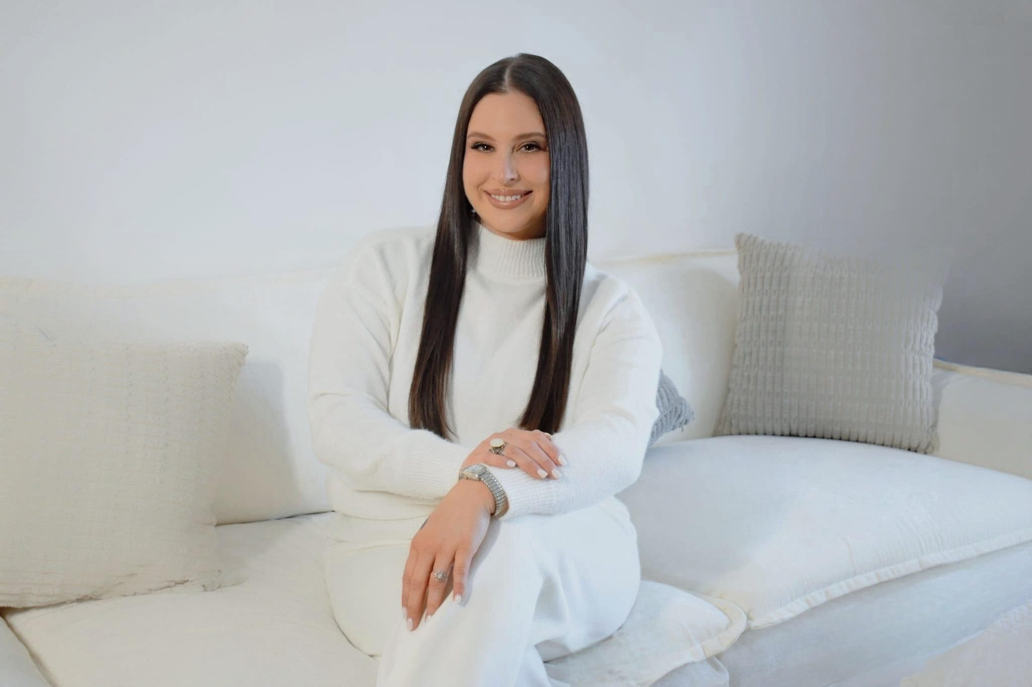 A woman sitting on a white sofa, smiling, wearing a white sweater and white pants, with her arms crossed on her lap, in a bright room with neutral decor.