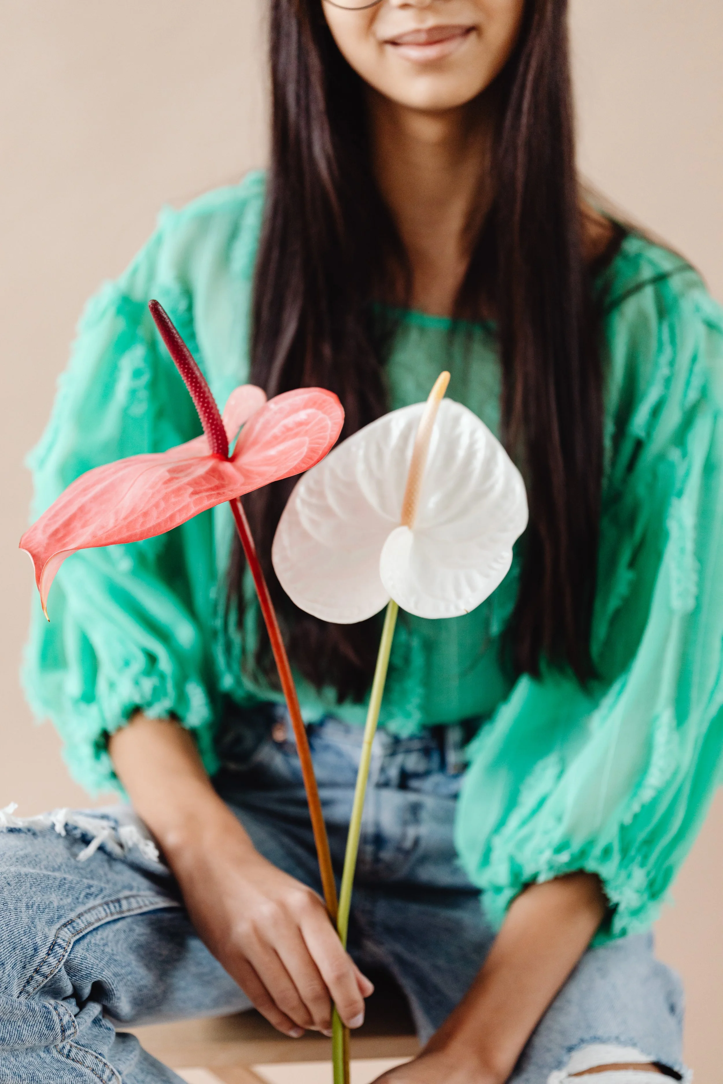 A woman with long dark hair, wearing a green top, holding three colorful artificial anthurium flowers, pink, white, and white with a slight pink hue, in front of her.