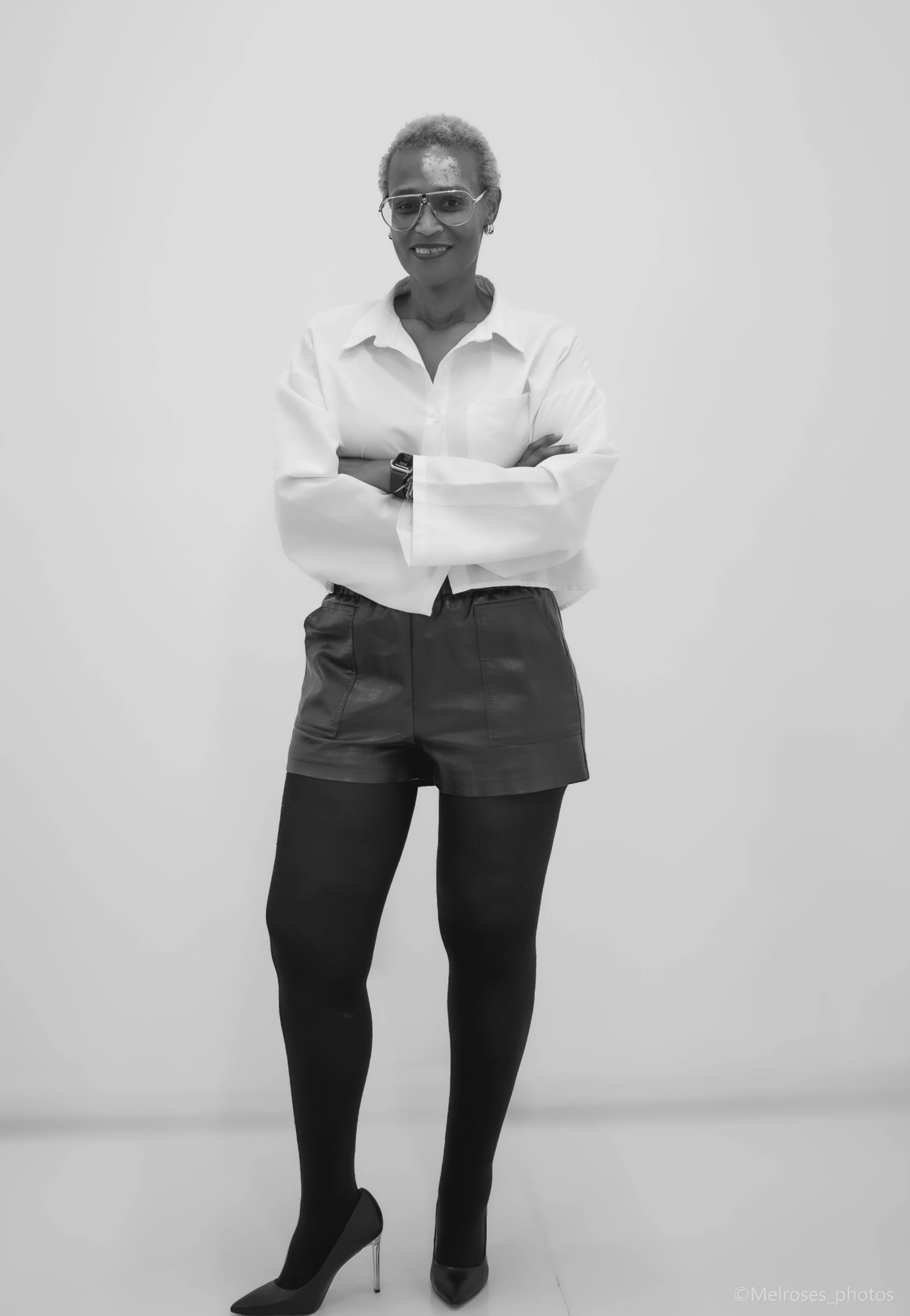 A confident woman with short blond hair, wearing glasses, a white button-up shirt, leather shorts, black tights, and high heels, standing against a plain background with her arms crossed.