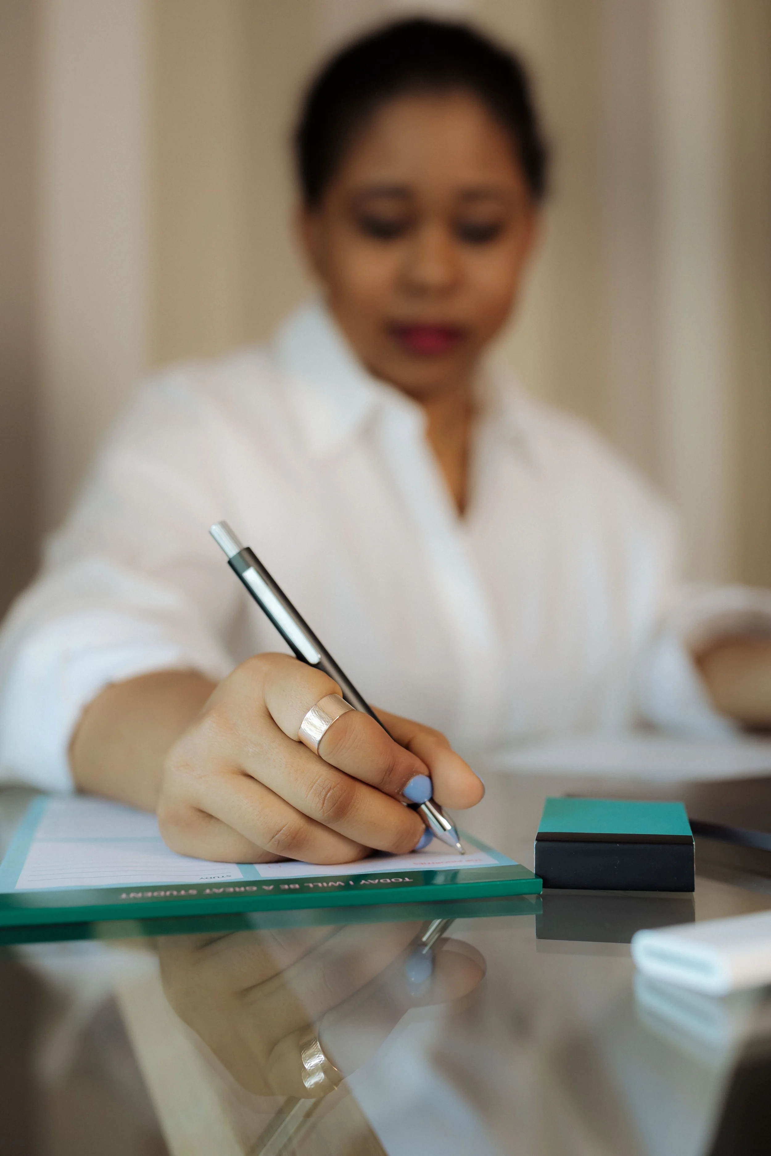 A woman in a white shirt writing on a green notepad with a pen, with a blurred focus on her face and sheet of paper, sitting at a reflective glass table.