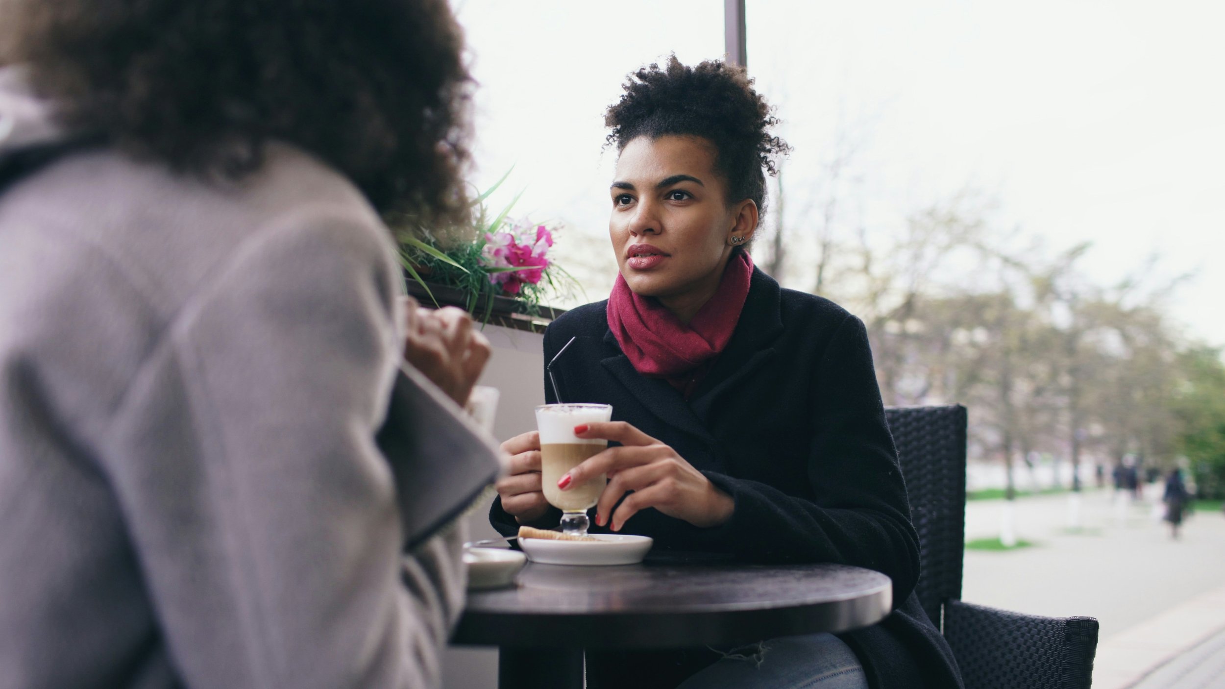 Two women having a conversation at a coffee shop, one holding a latte with whipped cream, both sitting at a small table near a window with a view of the outdoors.