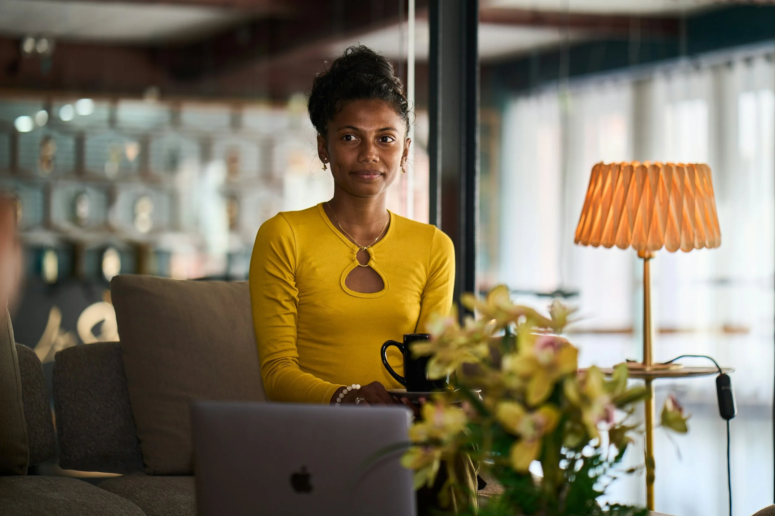 A woman in a yellow top sitting on a sofa in a well-lit room, holding a coffee cup, with a laptop and a flower arrangement in front of her, and a lamp to her side.