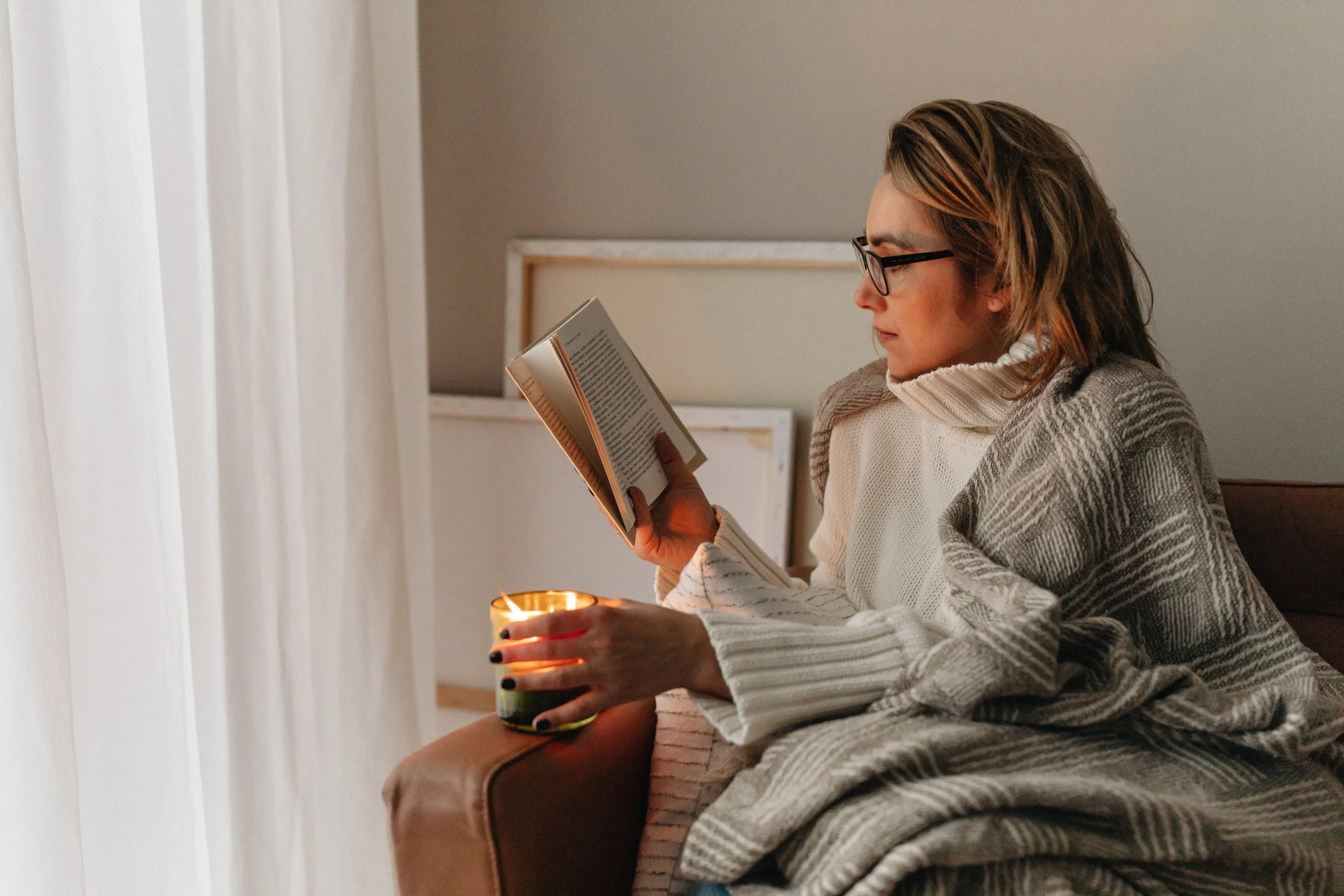 Woman sitting on a couch, reading a book and holding a lit candle.