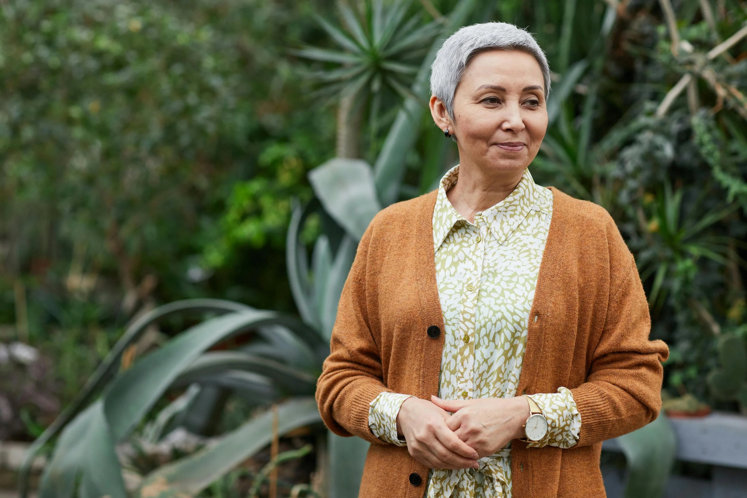 A woman with short gray hair standing outdoors among green plants, wearing a brown cardigan over a patterned shirt, and looking to the side.