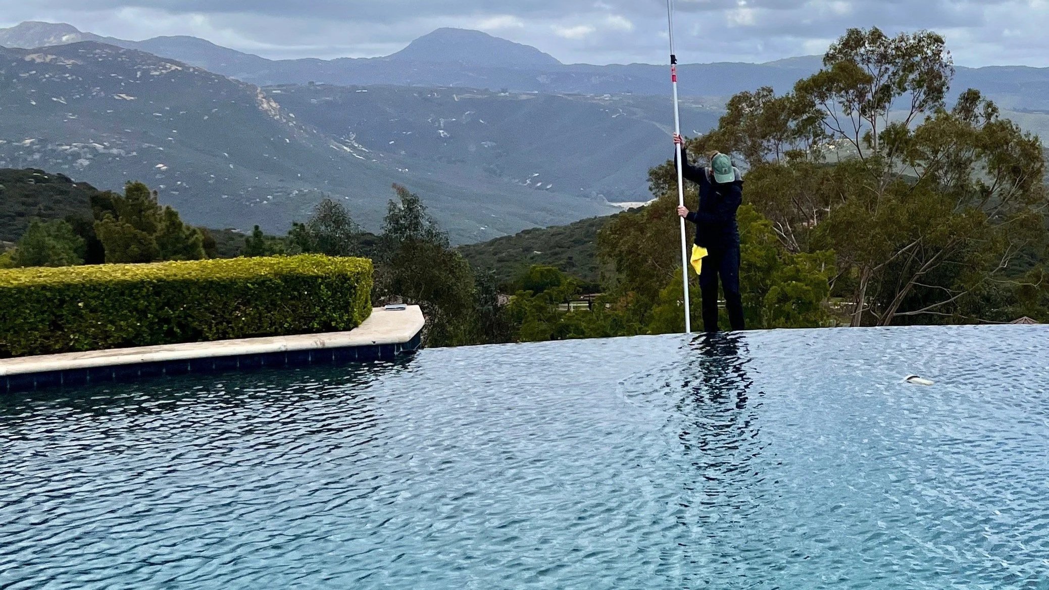 Person cleaning the edge of a swimming pool with a pole, greenery and mountains in the background.
