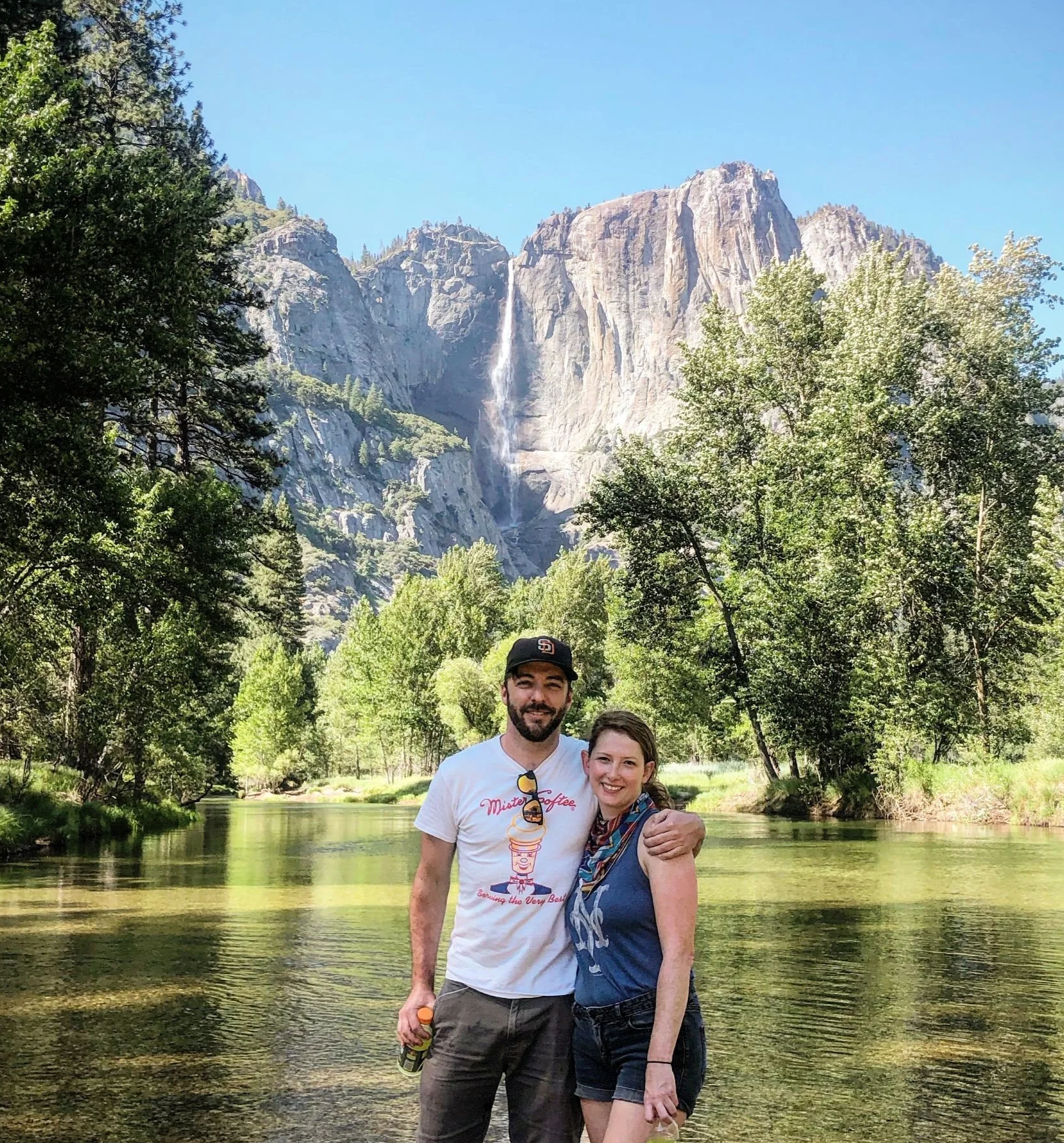 A couple standing together smiling in front of a river with trees, mountains, and a waterfall in the background.