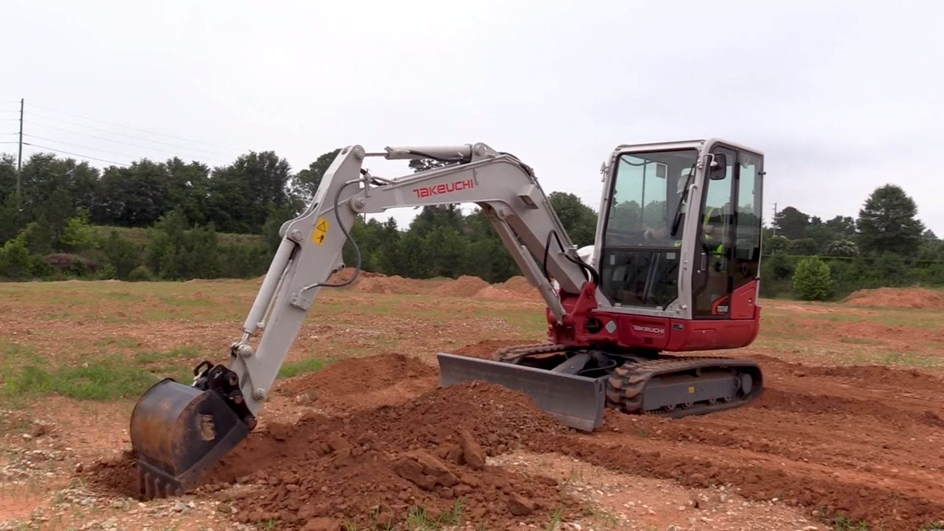A small red and gray Takeuchi mini excavator digging into the reddish soil on a construction site.