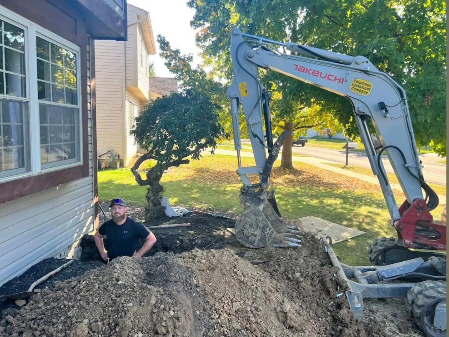 Man working at a construction site digging near a house with a small, twisted tree and a large excavator.