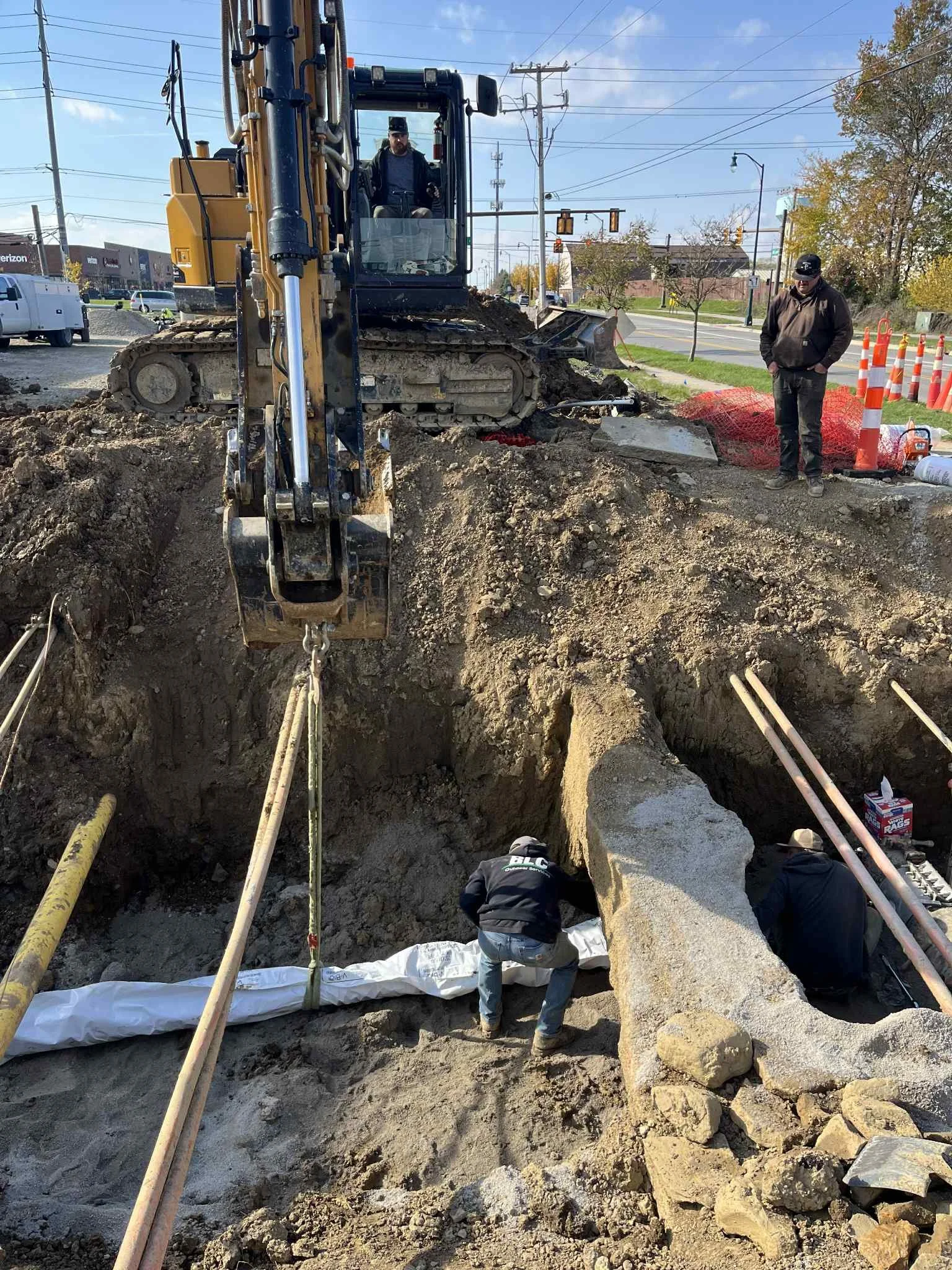 A construction site with an excavator digging a large trench on a city street. Two workers are inside the trench working on underground pipes, while another worker stands outside observing. Traffic and buildings are visible in the background.