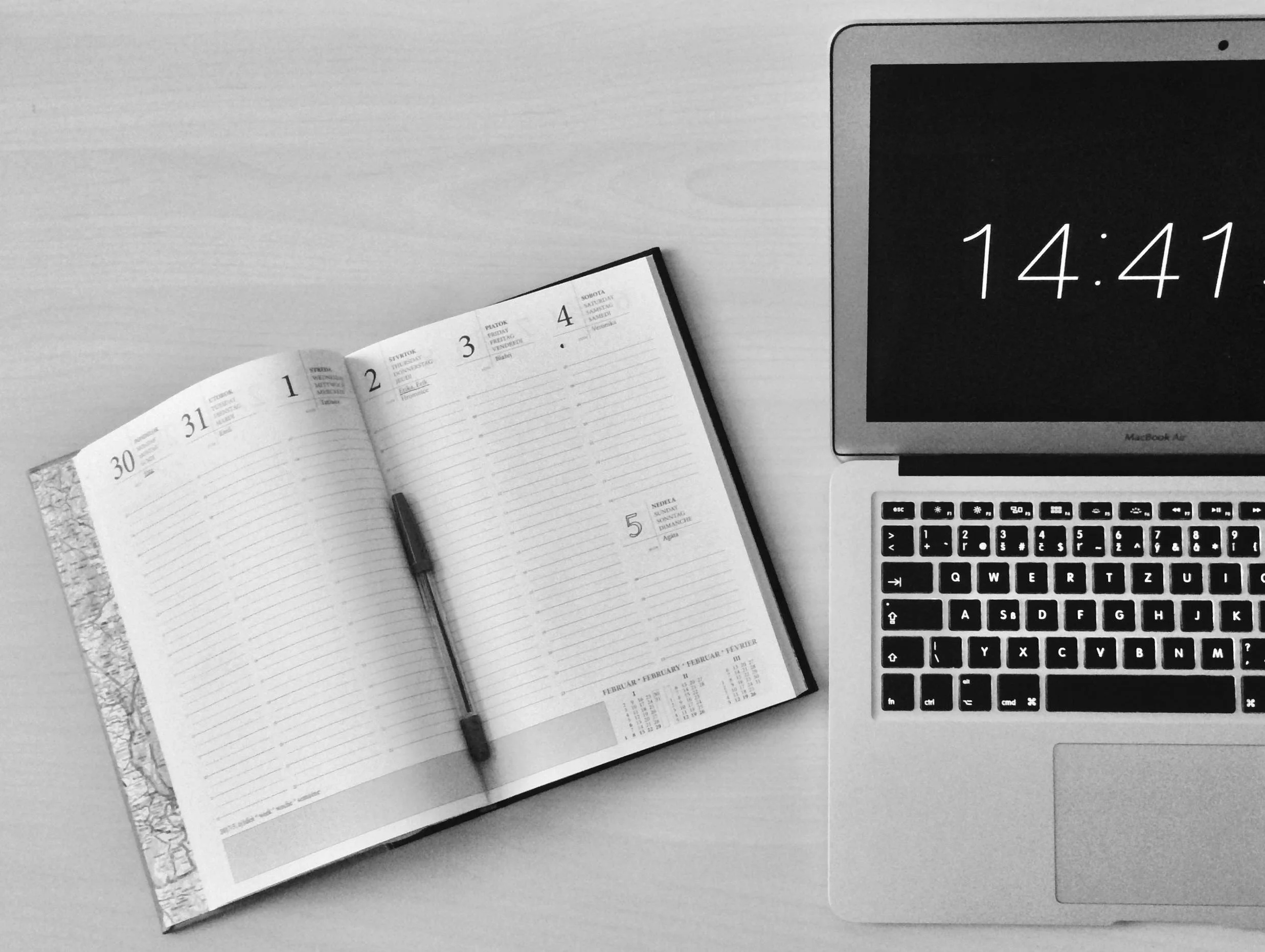Open weekly planner with a pen on left and a MacBook Air laptop with a digital clock display on the right on a light-colored desk.
